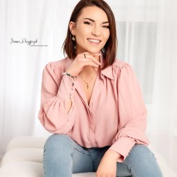 Joanna Skrzypczak Fotografia - Uśmiechnięta brunetka w różowej koszuli i jeansach siedzi na białej sofie, opierając podbródek na dłoni, z widocznym logo fotografa w lewym górnym rogu.