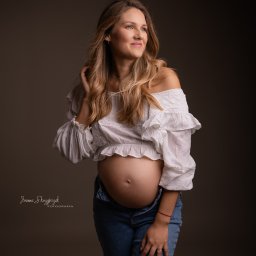 Joanna Skrzypczak Fotografia - Portret kobiety w ciąży, w białej bluzce z falbanami i rozpiętych dżinsach, z ręką we włosach, na ciemnym tle.