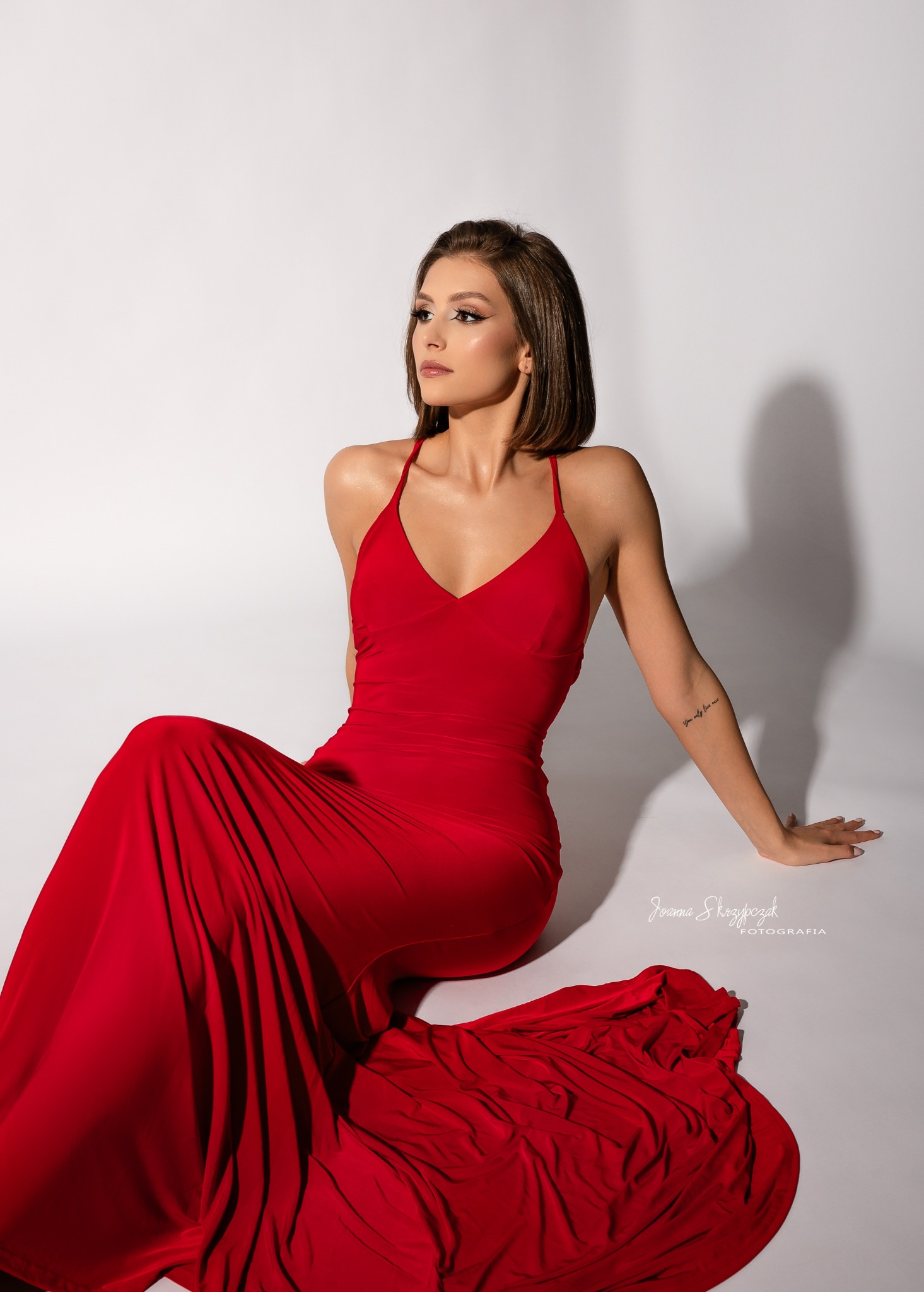 Portret kobiety w czerwonej, satynowej sukni z odkrytymi plecami, siedzącej na białym tle, z delikatnym makijażem i krótkimi brązowymi włosami, spoglądającej w bok, z widocznym tatuażem na ramieniu.