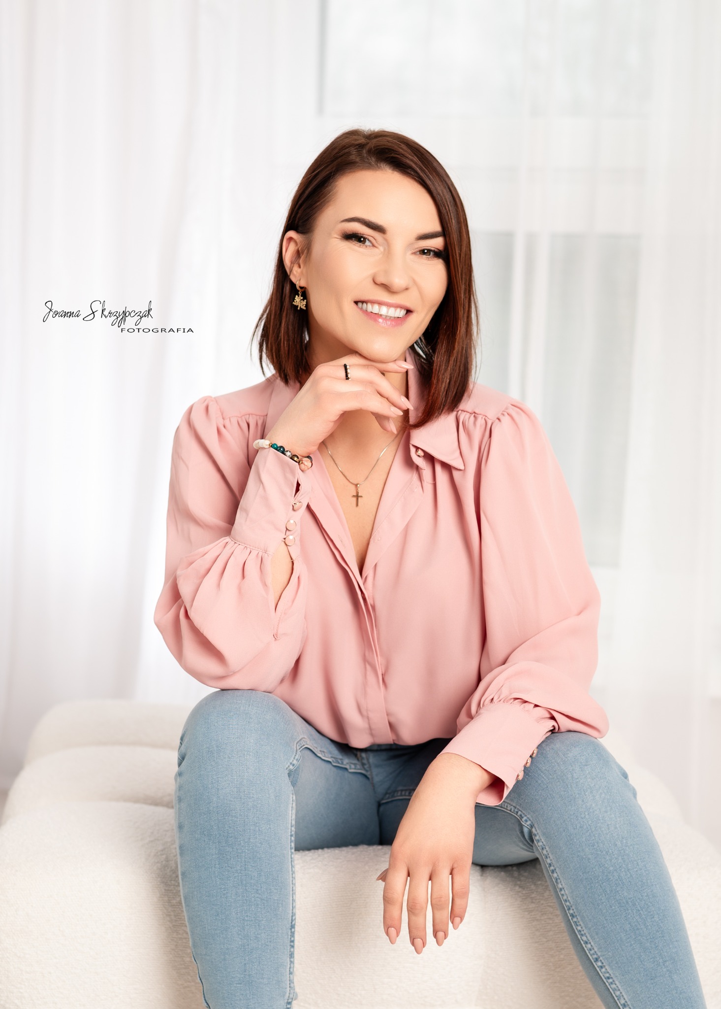 Uśmiechnięta brunetka w różowej koszuli i jeansach siedzi na białej sofie, opierając podbródek na dłoni, z widocznym logo fotografa w lewym górnym rogu.