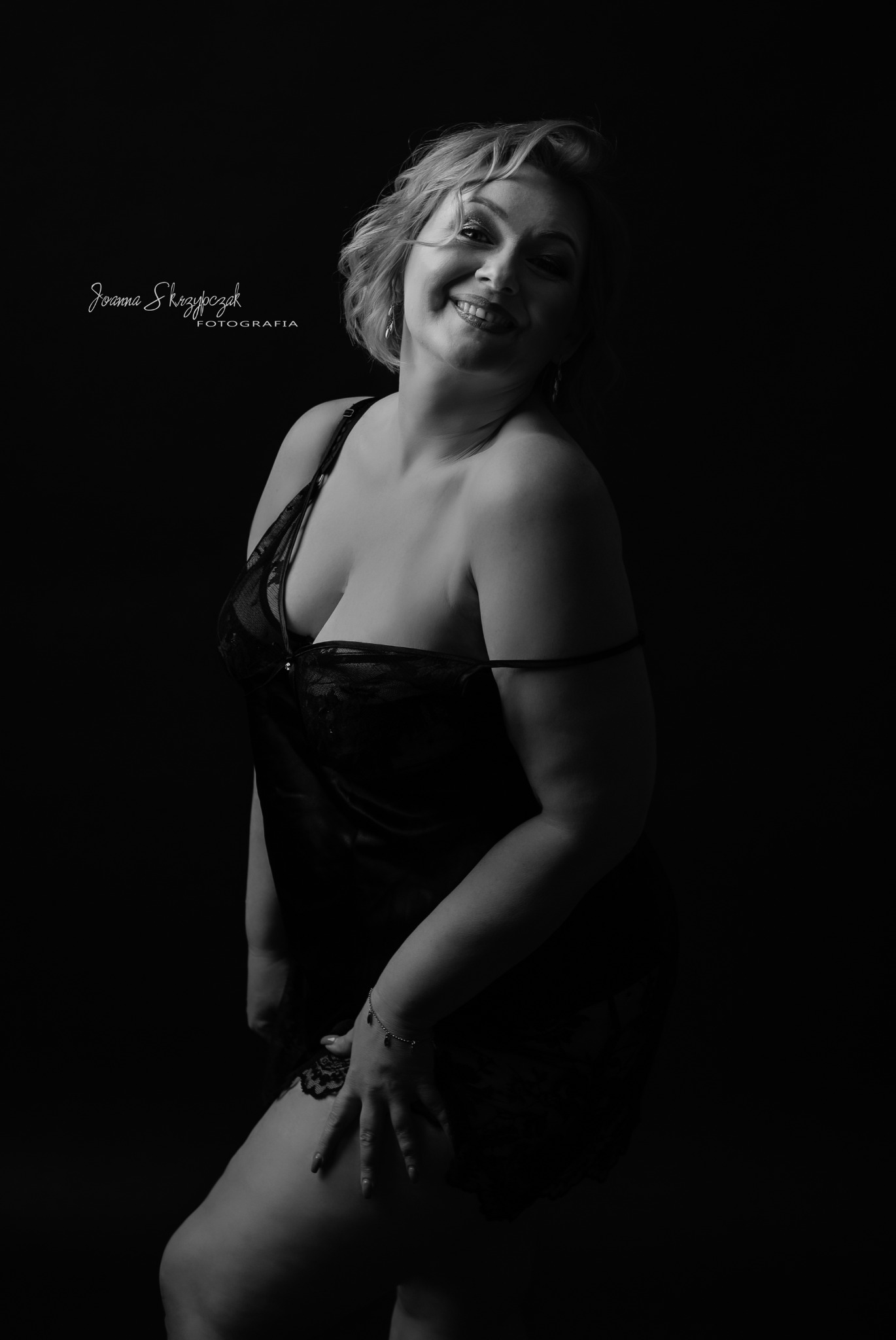 Portret kobiety w czarnej bieliźnie na ciemnym tle, z delikatnym uśmiechem i rozpuszczonymi blond włosami, uchwycony w klimatycznym, czarno-białym ujęciu.