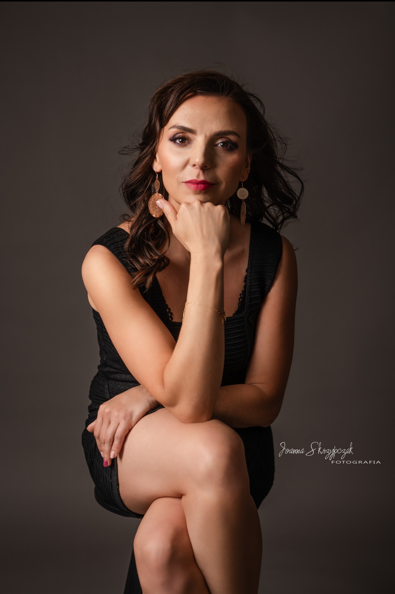 Portret kobiety 40+ w czarnej sukience, z brązowymi włosami i złotymi kolczykami, pozującej na szarym tle z ręką pod brodą.