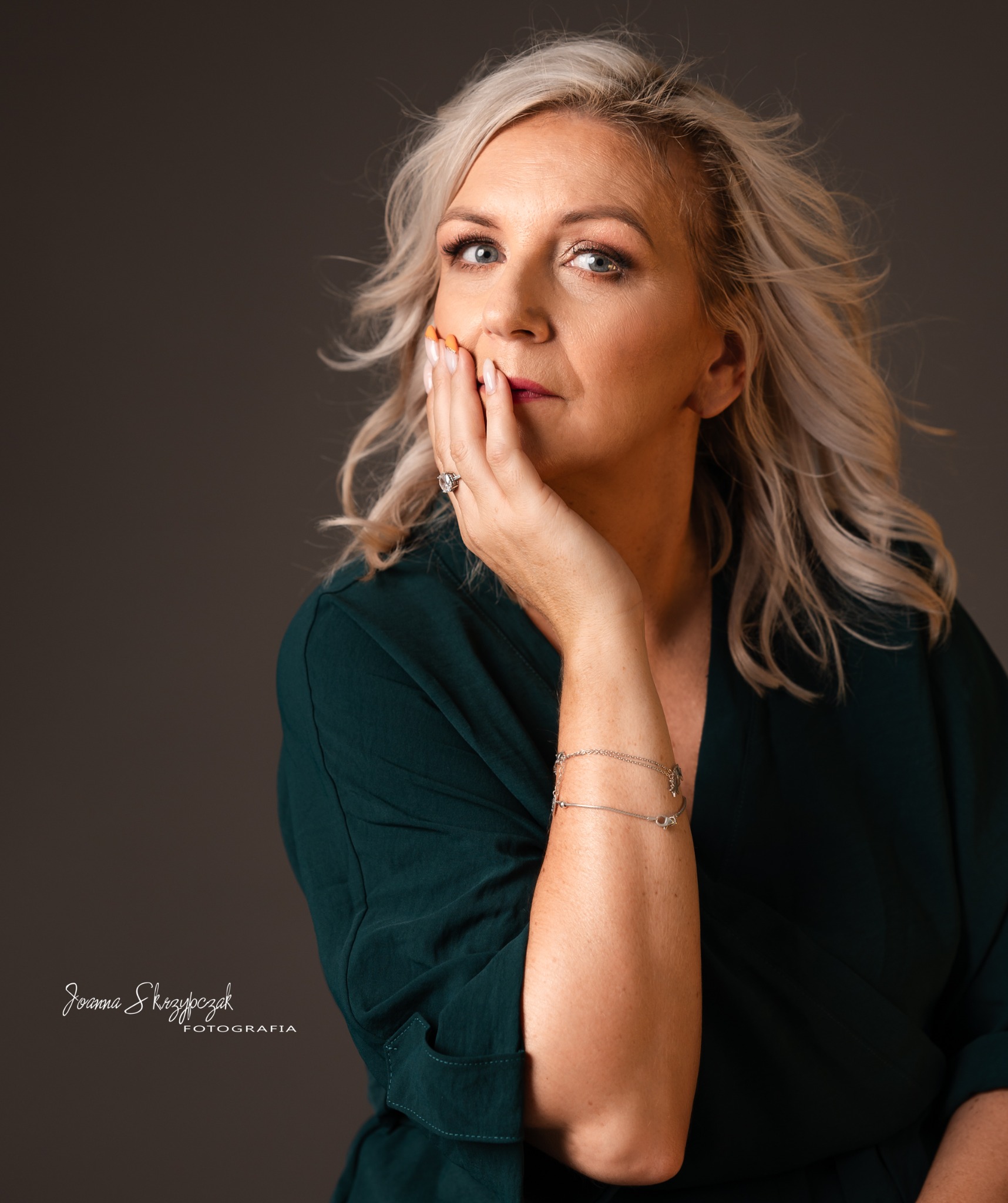 Portret kobiety z blond włosami w zielonej sukience, dłoń przy twarzy, delikatny makijaż i biżuteria.