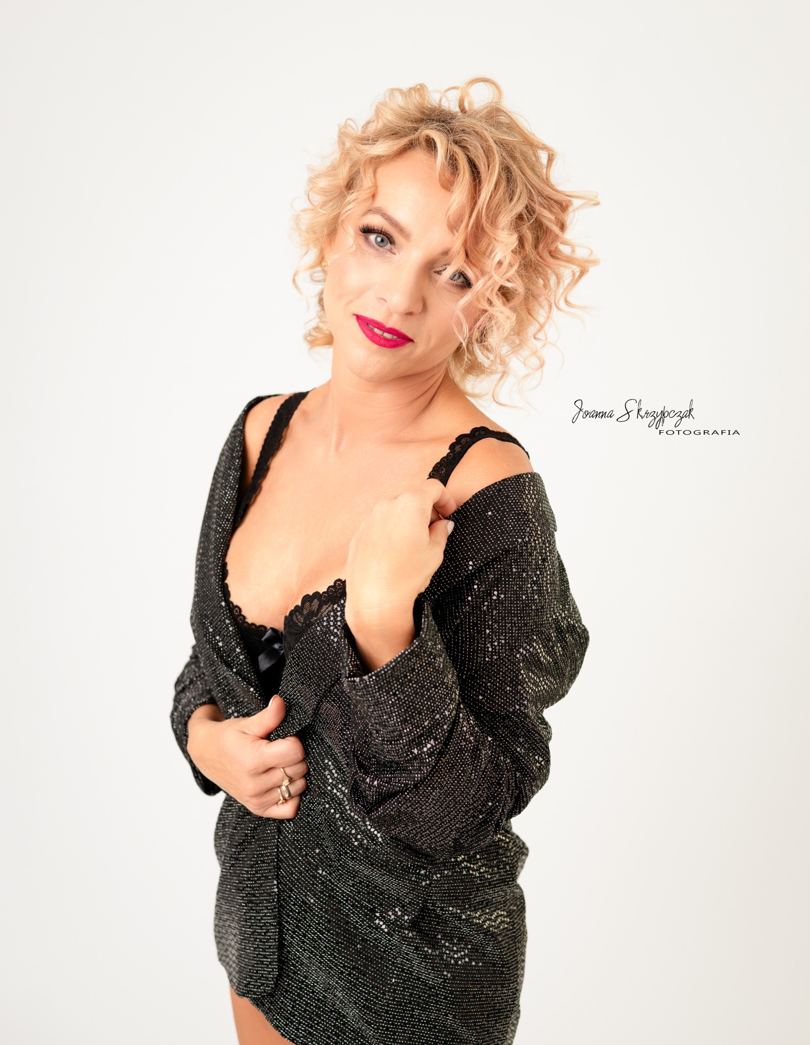 Kobieta z blond lokami, w czarnej, błyszczącej marynarce i czarnej bieliźnie, pozuje na białym tle z delikatnym uśmiechem i czerwonymi ustami.