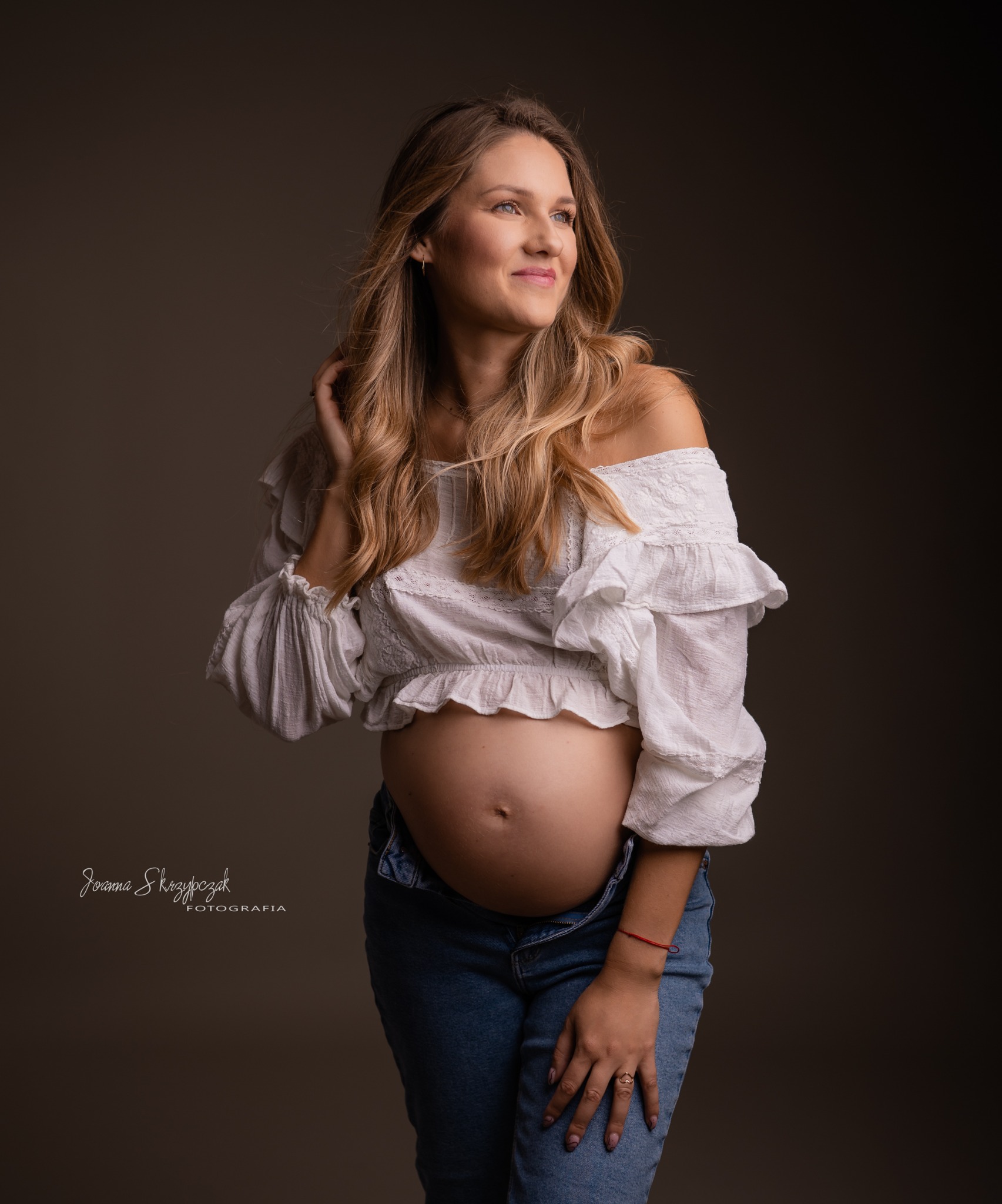 Portret kobiety w ciąży, w białej bluzce z falbanami i rozpiętych dżinsach, z ręką we włosach, na ciemnym tle.