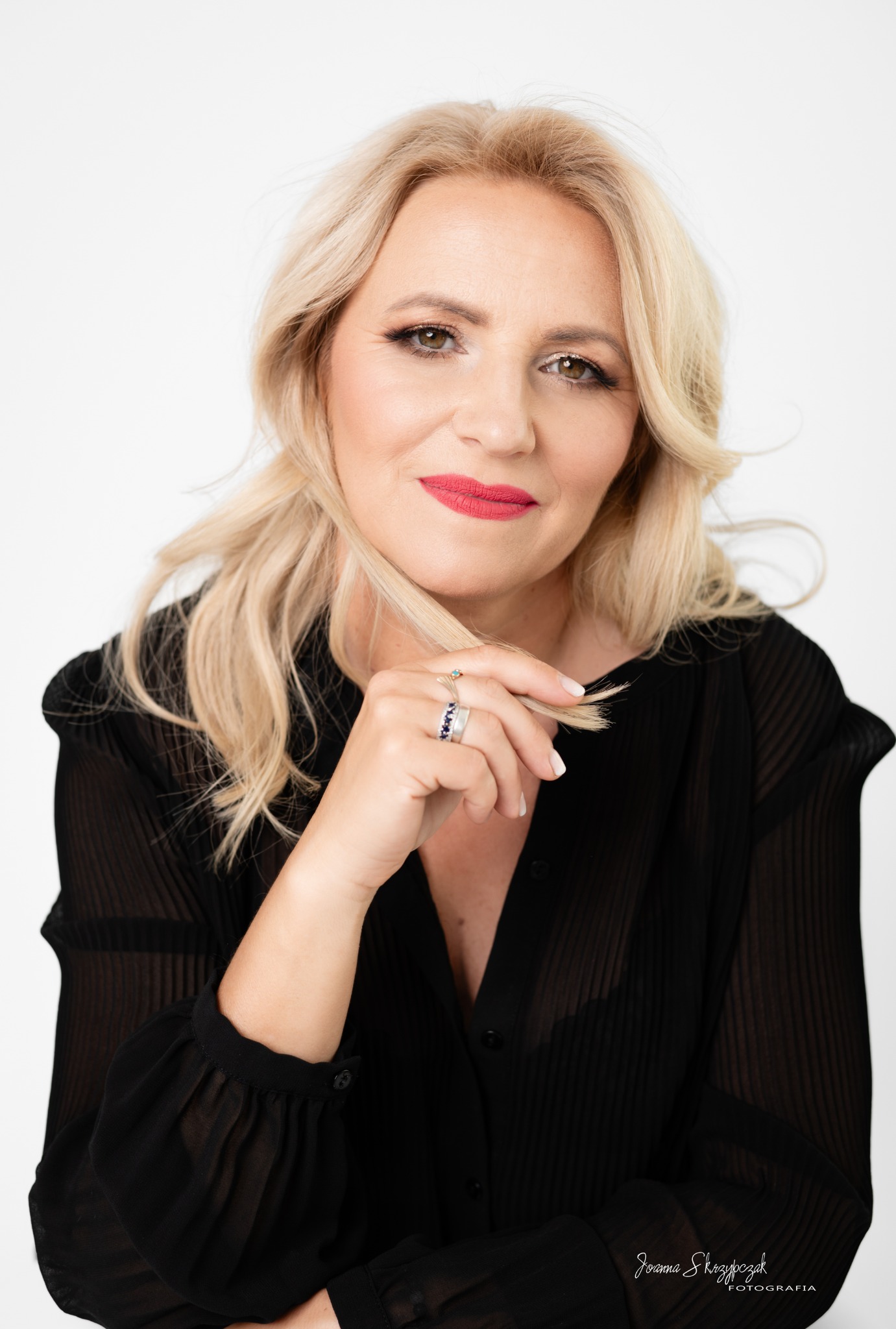 Portret kobiety z blond włosami, w czarnej bluzce, z delikatnym makijażem i pierścionkami na palcu, trzymającej pasmo włosów na białym tle.