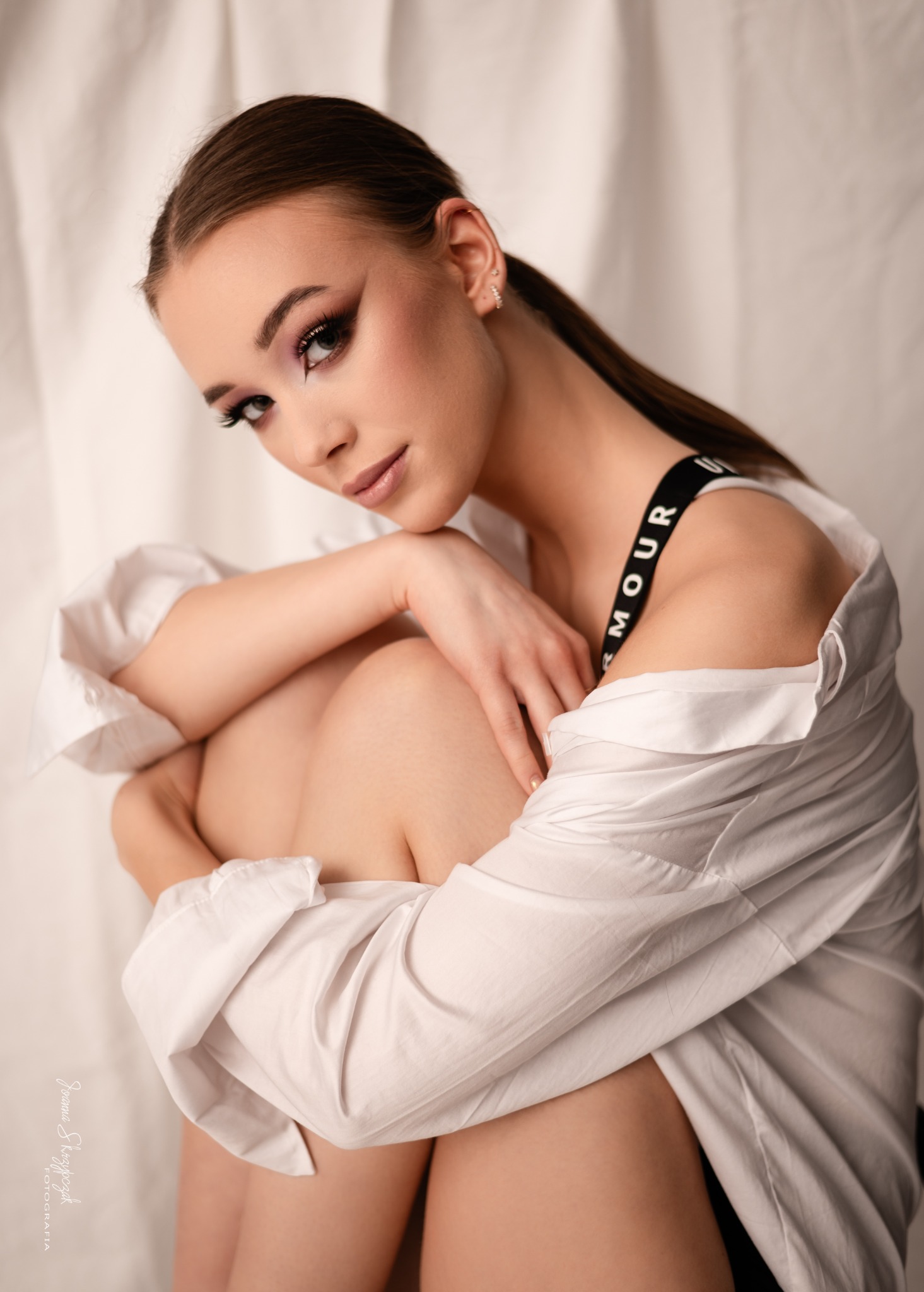 Portret młodej kobiety z profesjonalnym makijażem, ubrana w białą koszulę, obejmująca kolana na tle białego materiału.