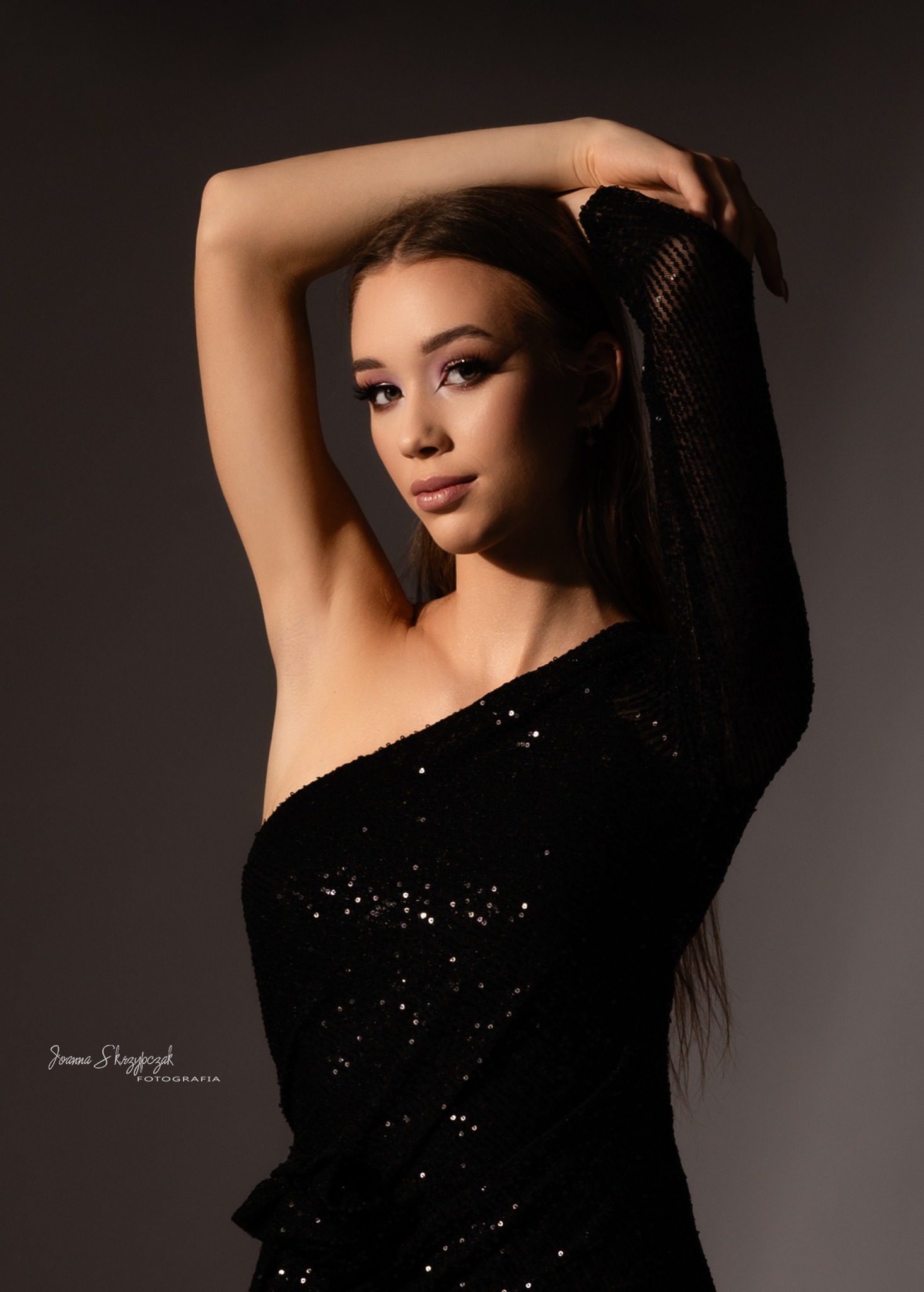 Portret kobiety w czarnej, cekinowej sukience na jedno ramię, z ręką uniesioną nad głową, w studyjnym oświetleniu.