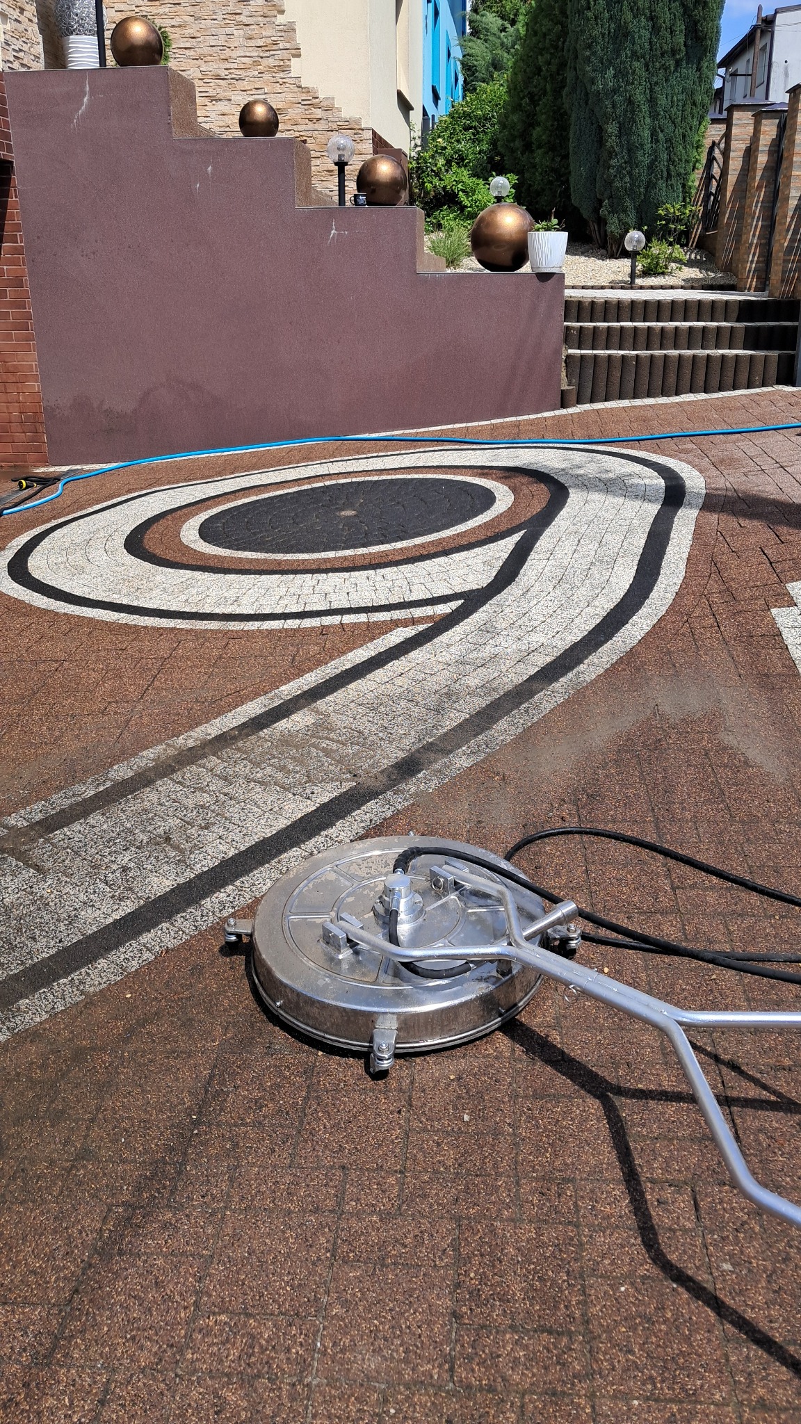 Okrągła myjka ciśnieniowa w trakcie czyszczenia wzorzystej kostki brukowej na podjeździe; widoczne ciemne i jasne pasy czyszczonej powierzchni.