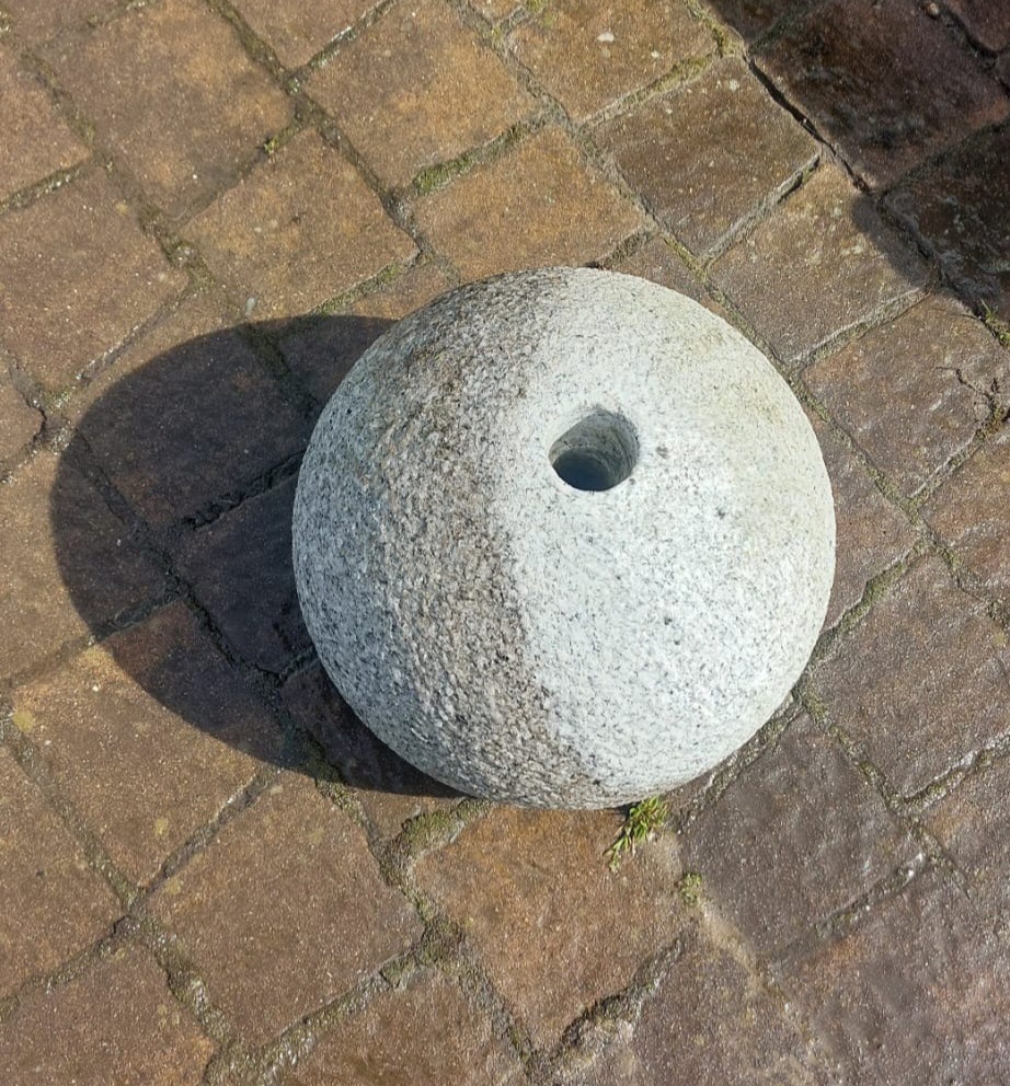 Granitowa kula z otworem, częściowo oczyszczona, leżąca na brązowej kostce brukowej.