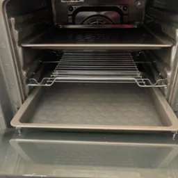 Wnętrze czystego piekarnika z blachami i rusztem, widok z przodu na otwarte drzwiczki. Numeracja prowadnic na ściankach bocznych.