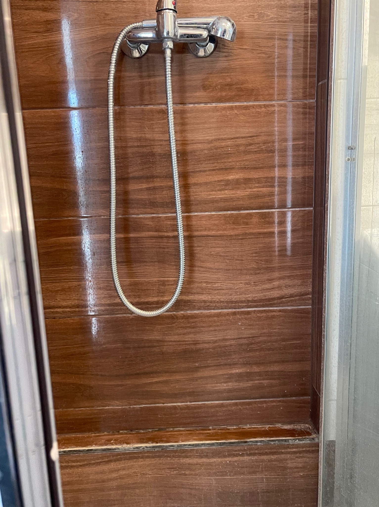 Błyszcząca, brązowa ściana kabiny prysznicowej z widoczną słuchawką i wężem prysznicowym. Widoczne ślady po wodzie na panelach.
