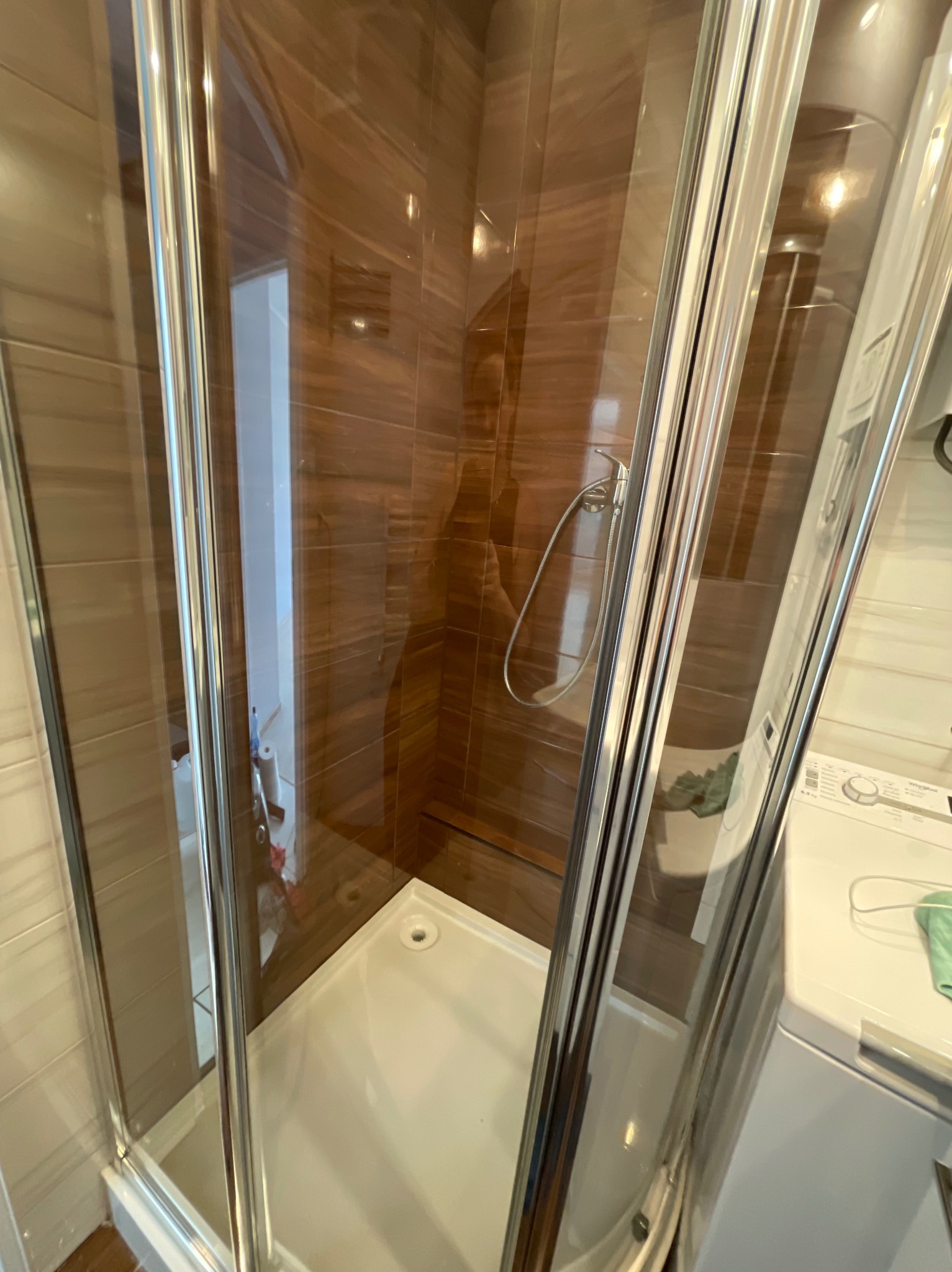 Szklana kabina prysznicowa z brązowymi płytkami imitującymi drewno w łazience z pralką. Widok z zewnątrz, czysta i gotowa do użytku.