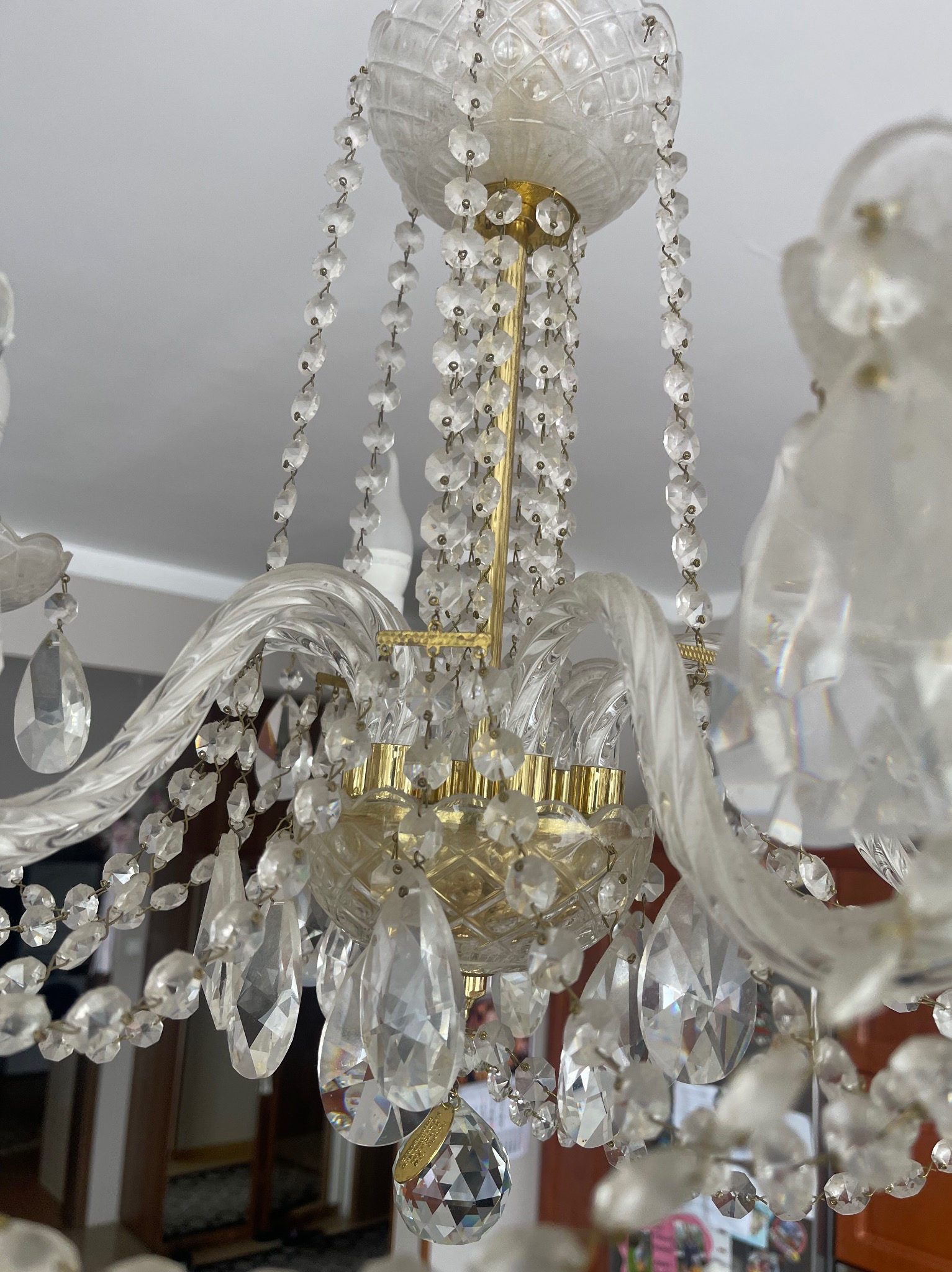 Kryształowy żyrandol z bliska, z licznymi szklanymi elementami i złotymi detalami, wiszący pod białym sufitem. Widoczne elementy wystroju wnętrza.