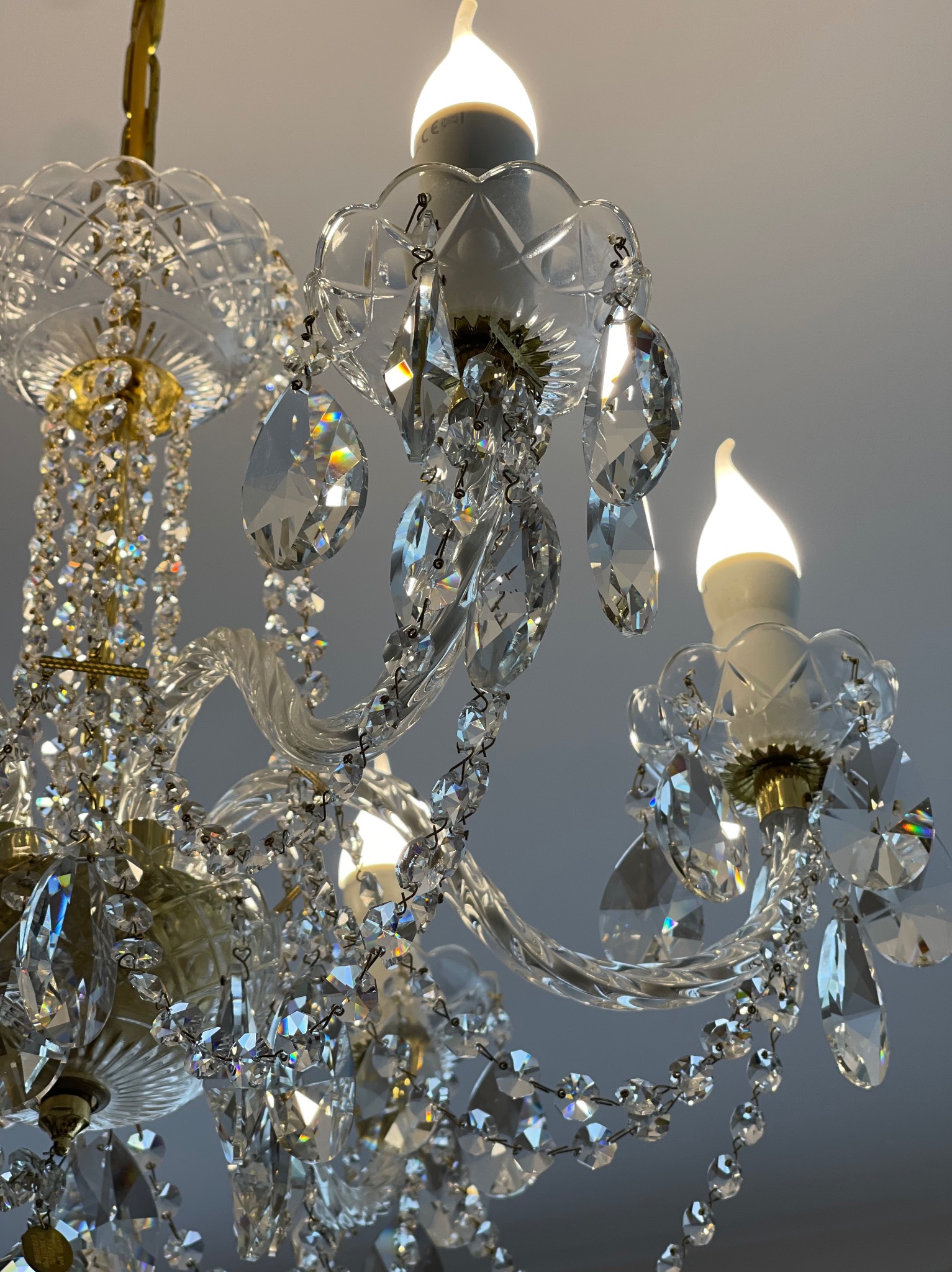 Kryształowy żyrandol z ozdobnymi łańcuchami i szklanymi elementami, oświetlony żarówkami imitującymi płomień świecy, widok z dołu na tle jasnego sufitu.