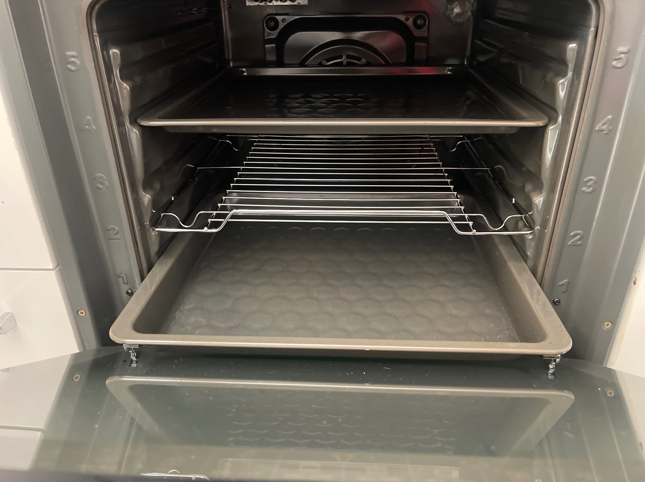 Wnętrze czystego piekarnika z blachami i rusztem, widok z przodu na otwarte drzwiczki. Numeracja prowadnic na ściankach bocznych.