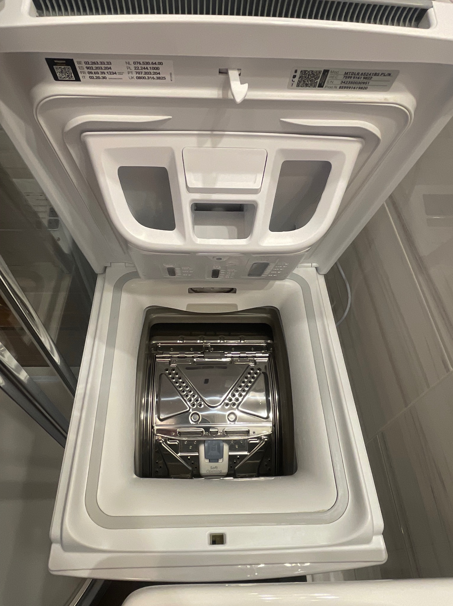 Biała pralka ładowana od góry z otwartym wiekiem, ukazująca wnętrze bębna ze stali nierdzewnej. Widoczne numery kontaktowe i model na wewnętrznej stronie wieka.