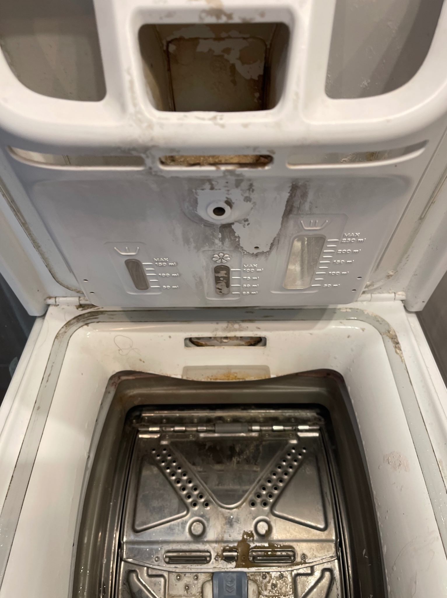 Z bliska wnętrze brudnej pralki z otwartą klapą, ukazujące osady i zabrudzenia wewnątrz bębna oraz na elementach plastikowych dozownika. Widoczne ślady użytkowania.