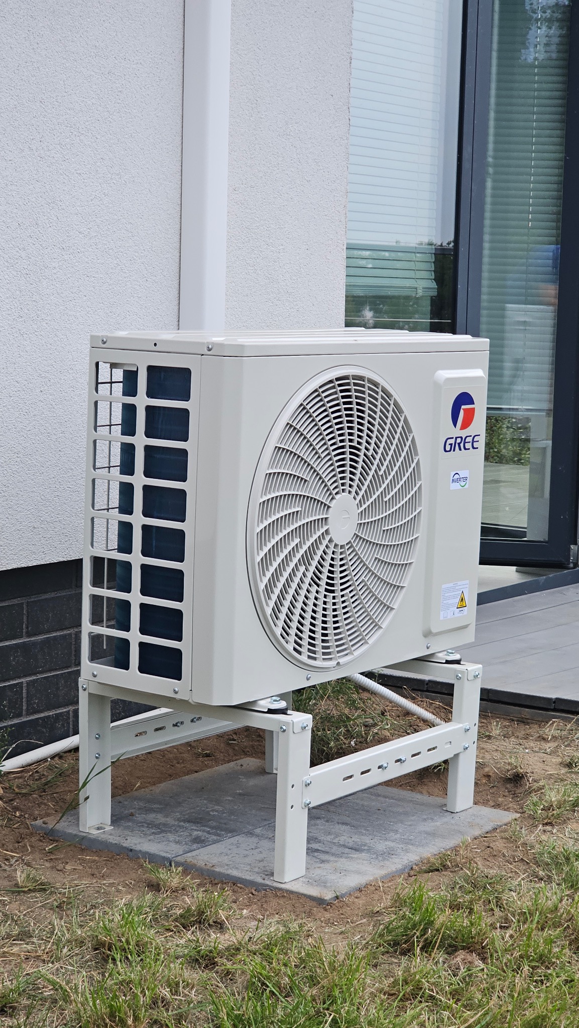 Zewnętrzna jednostka klimatyzacji GREE zamontowana na metalowym stelażu przy ścianie budynku z oknem.