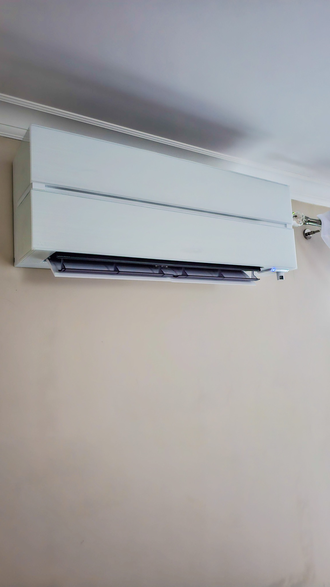 Biały, nowoczesny klimatyzator ścienny zamontowany w pokoju z beżową ścianą i białym sufitem z listwą dekoracyjną.