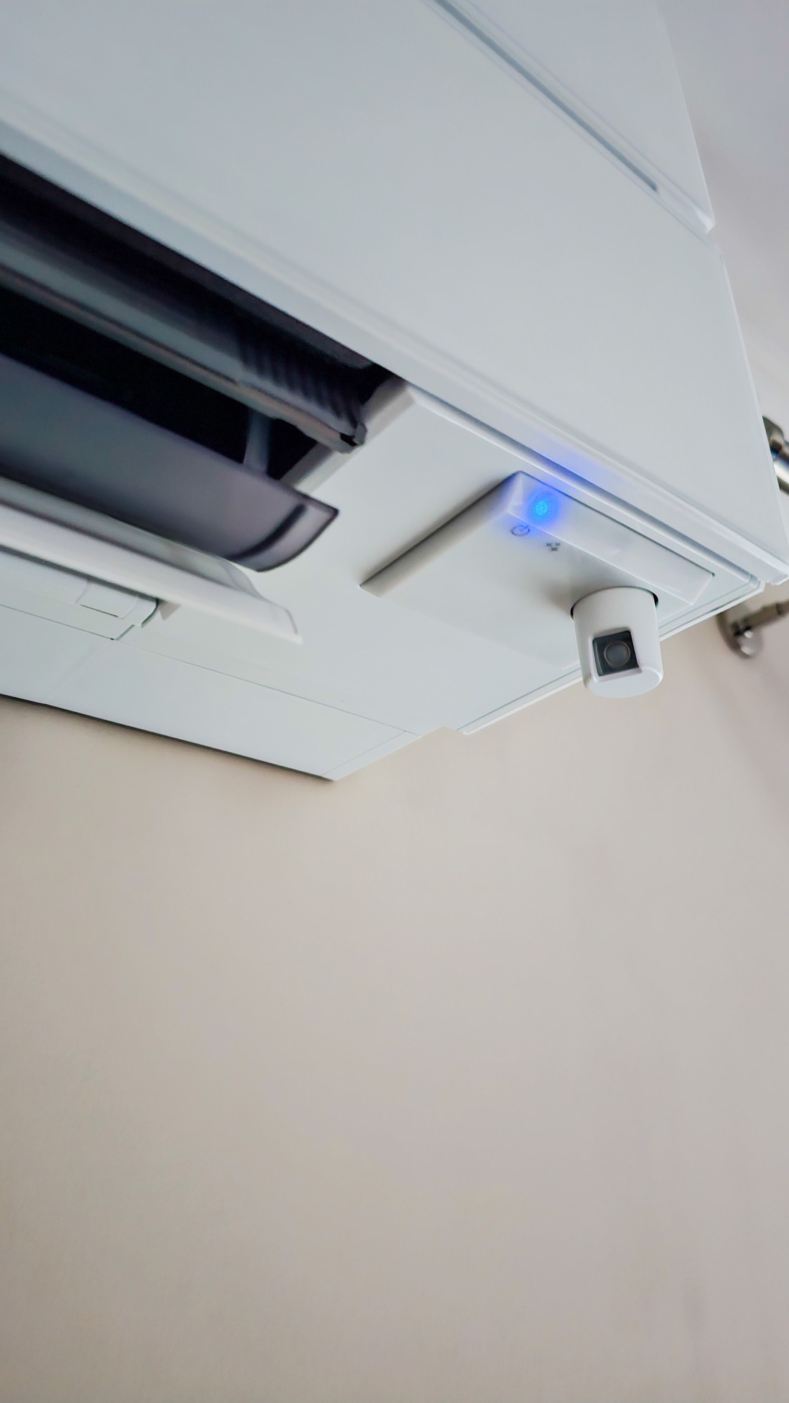 Nowoczesny, biały klimatyzator z widocznym czujnikiem ruchu i niebieską diodą LED, zamontowany na jasnej ścianie, z perspektywy dolnej.