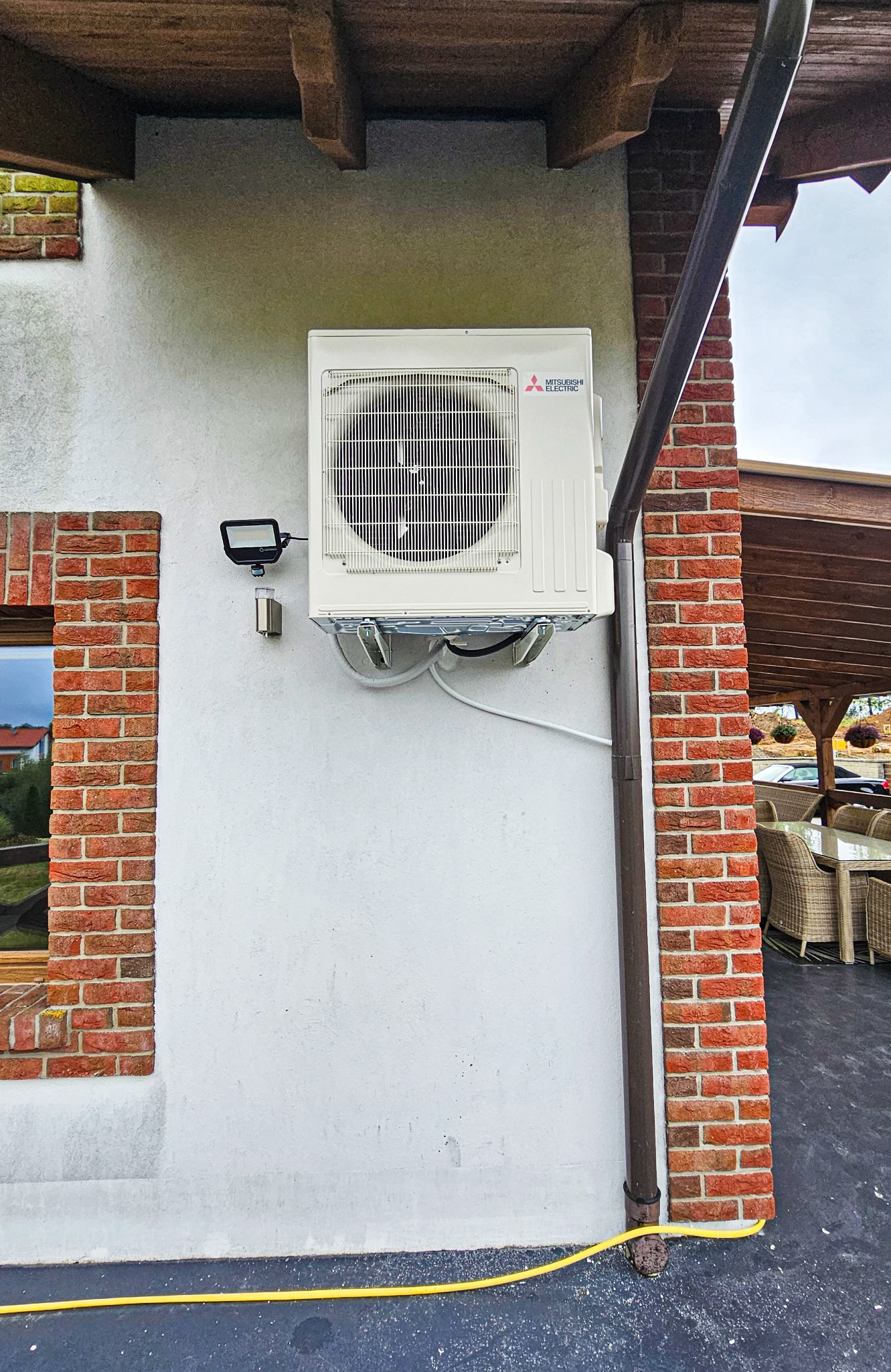 Biała jednostka zewnętrzna klimatyzacji Mitsubishi Electric zamontowana na elewacji domu, widoczny fragment rynny i okna z ceglaną obudową.