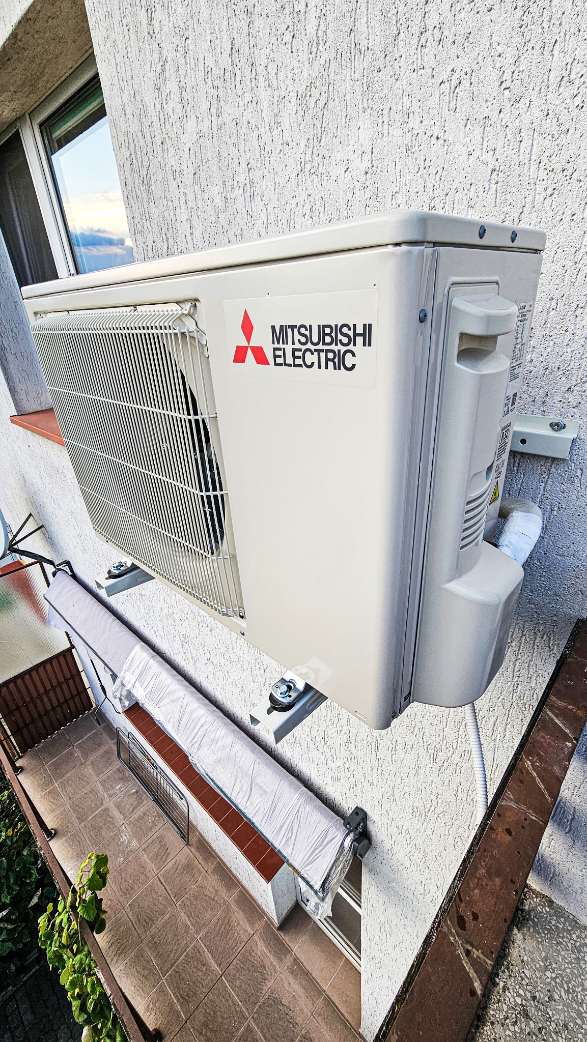 Zewnętrzna jednostka klimatyzacji Mitsubishi Electric zamontowana na elewacji budynku z widocznym balkonem i fragmentem okna, ujęcie z dołu.