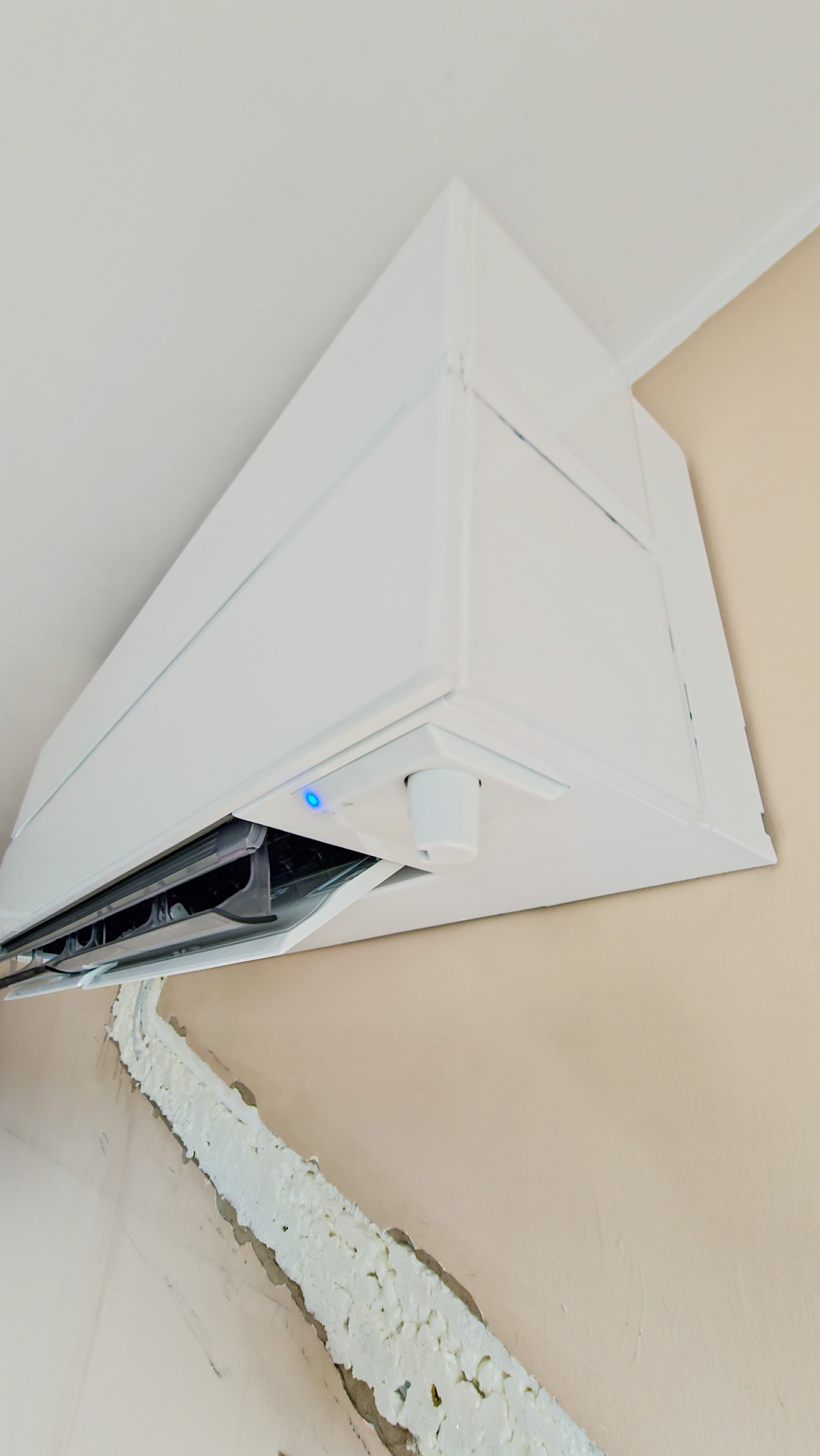 Biała jednostka wewnętrzna klimatyzatora Mitsubishi z niebieską diodą, zamontowana na ścianie z widoczną pianką montażową wokół przewodów.