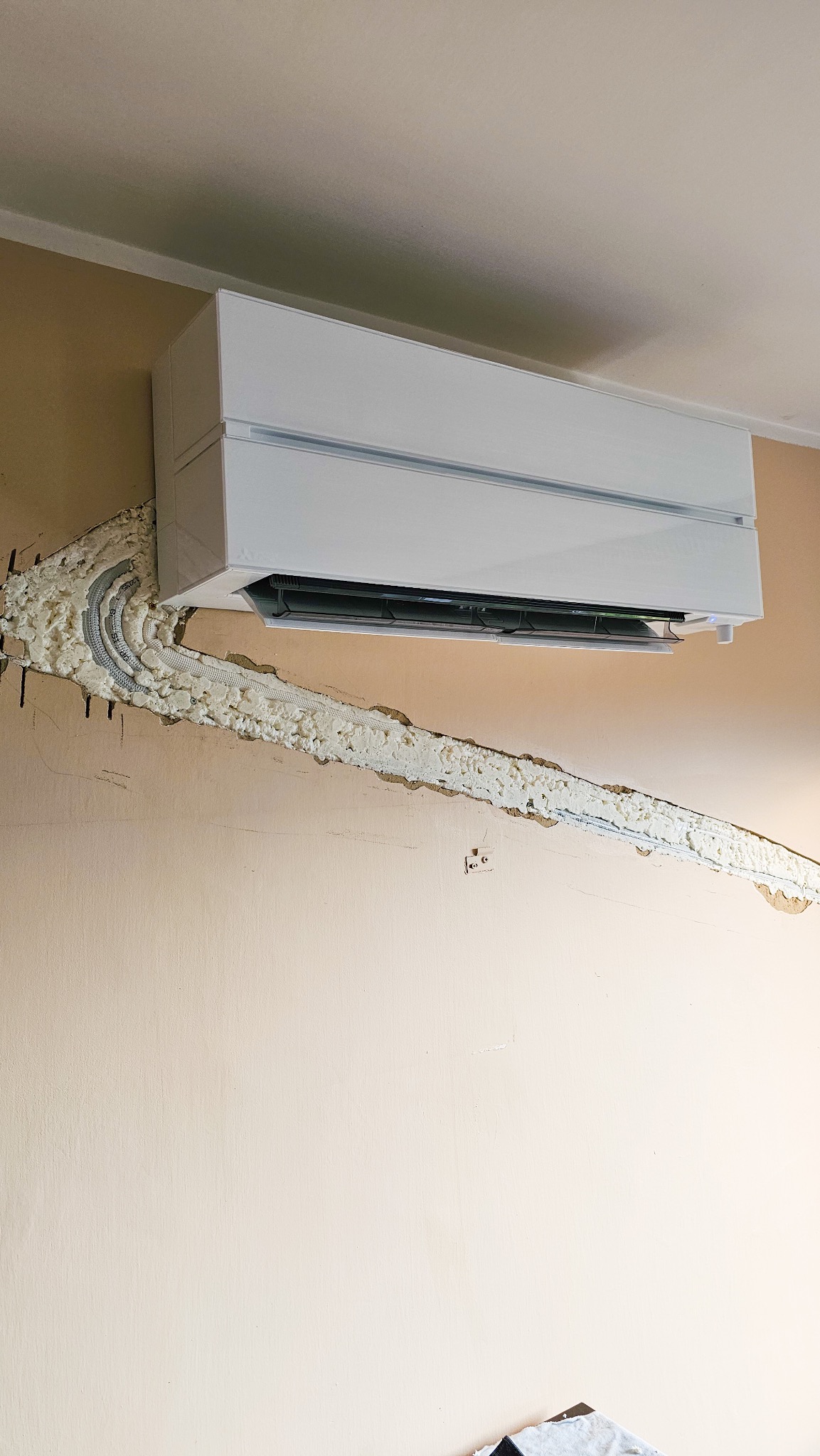 Biały klimatyzator Mitsubishi zamontowany na beżowej ścianie, z widocznymi przewodami zabezpieczonymi pianką montażową w bruździe. Widok z dołu, ukazujący etap instalacji.