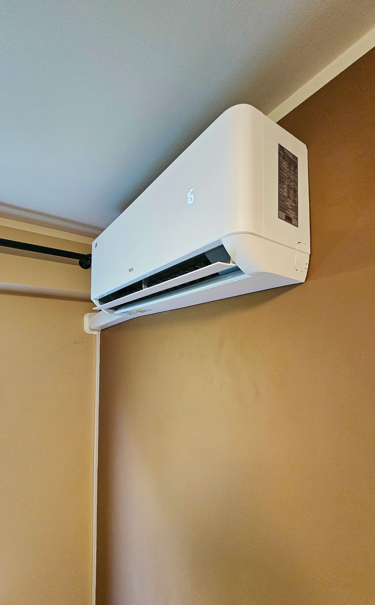 Nowoczesny, biały klimatyzator TCL zamontowany na ścianie w pomieszczeniu o beżowych ścianach, z wyświetlaczem temperatury wskazującym 16 stopni Celsjusza.