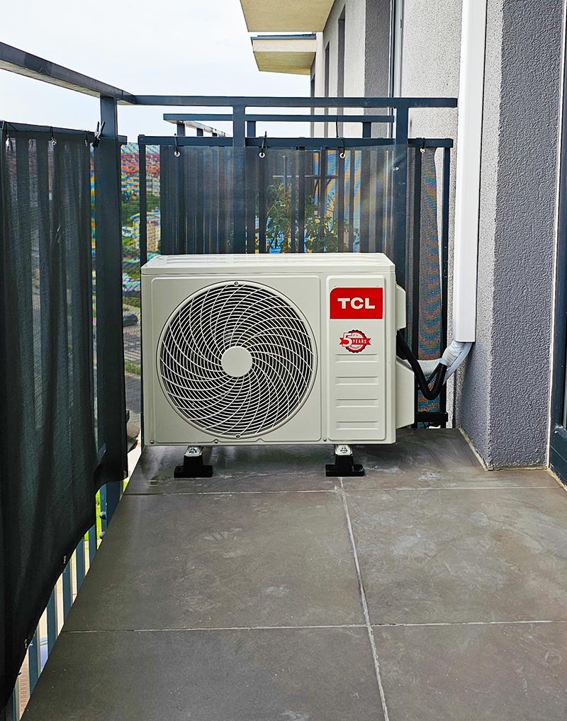 Zewnętrzna jednostka klimatyzacji TCL zamontowana na balkonie z szarą podłogą i czarną osłoną przeciwsłoneczną.