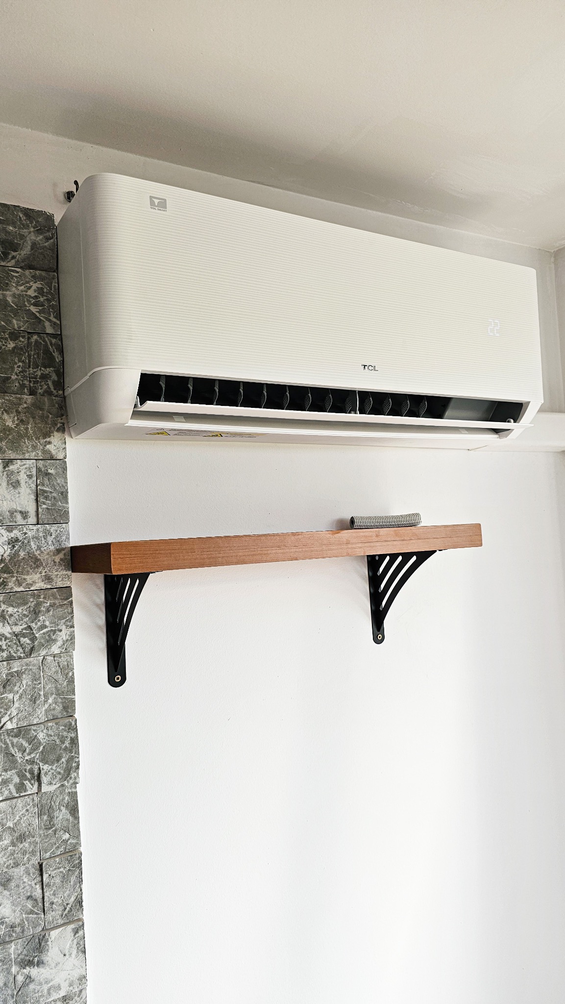 Biały klimatyzator TCL OCARINA TPG31 zamontowany na ścianie z dekoracyjną okładziną imitującą kamień, pod nim drewniana półka na czarnych wspornikach.