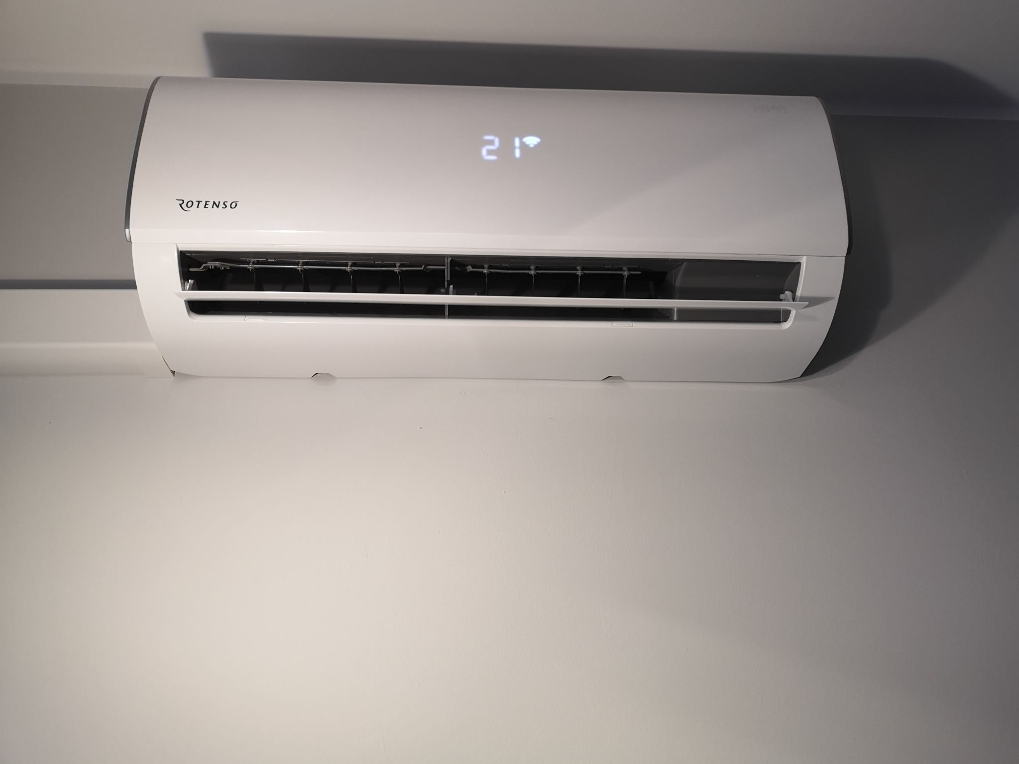 Biała jednostka klimatyzacyjna Rotenso z wyświetlaną temperaturą 21 stopni Celsjusza, zamontowana na szarej ścianie.