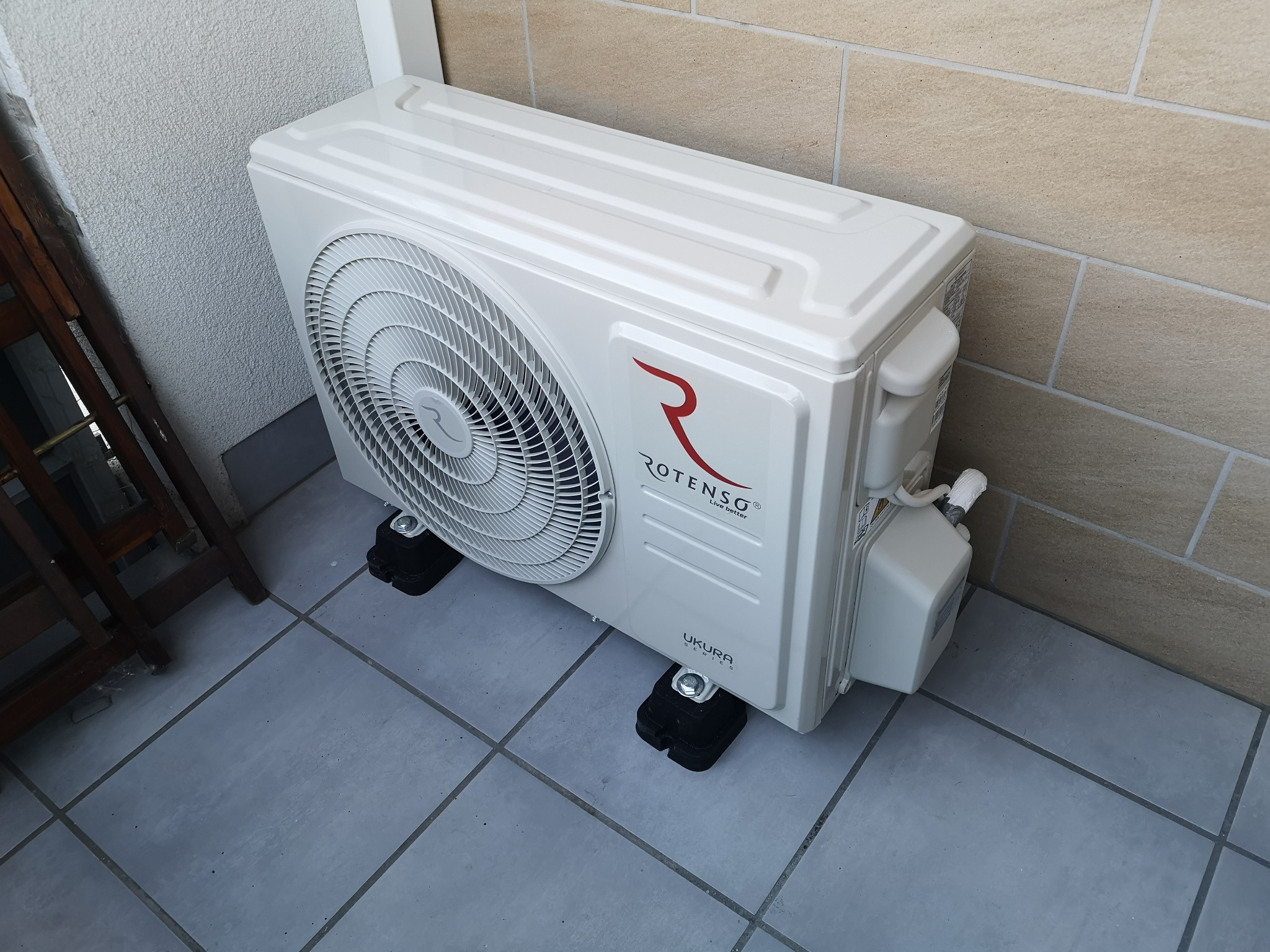 Zewnętrzna jednostka klimatyzacji Rotenso Ukura na czarnych podstawkach antywibracyjnych, zamontowana na szarych płytkach tarasu przy beżowej ścianie.