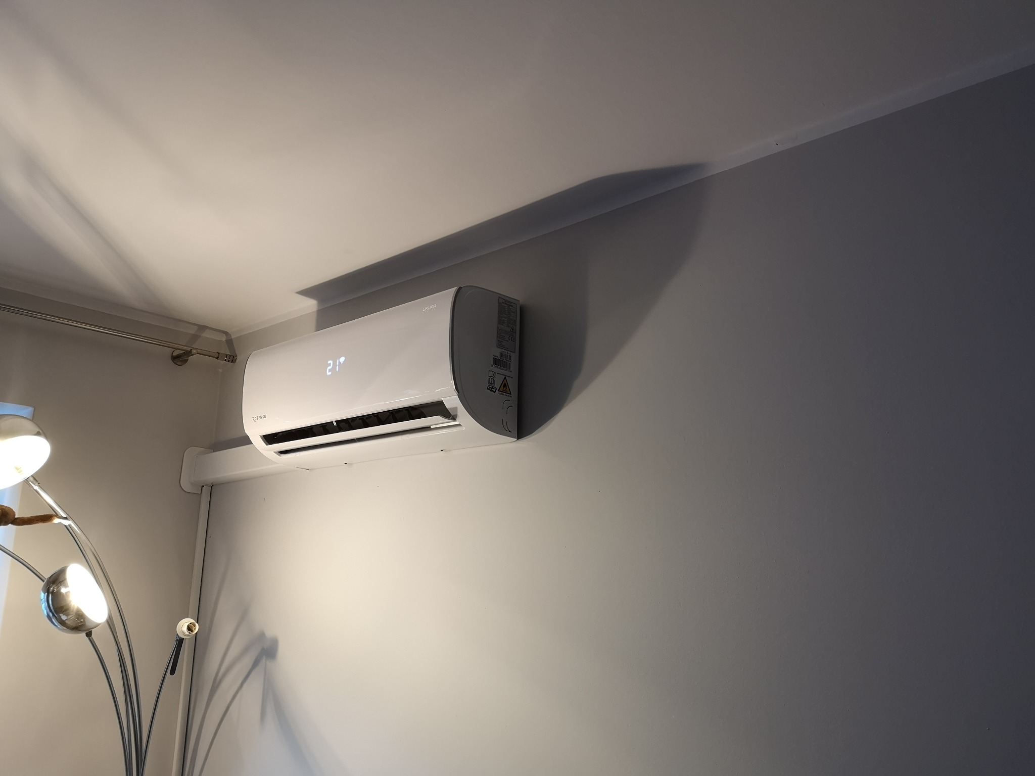 Biały klimatyzator ścienny z wyświetlaczem temperatury 21 stopni, zamontowany w jasnym pomieszczeniu z lampą podłogową.