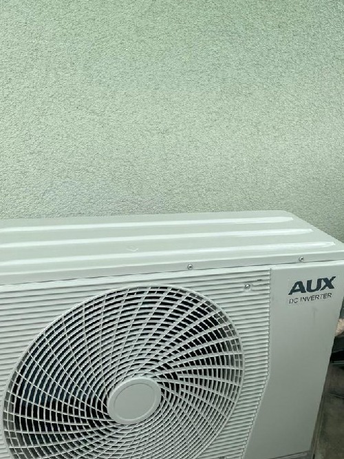 Zewnętrzna jednostka klimatyzacji AUX DC Inverter zamontowana na ścianie o szorstkiej fakturze, widoczne śruby mocujące obudowę.