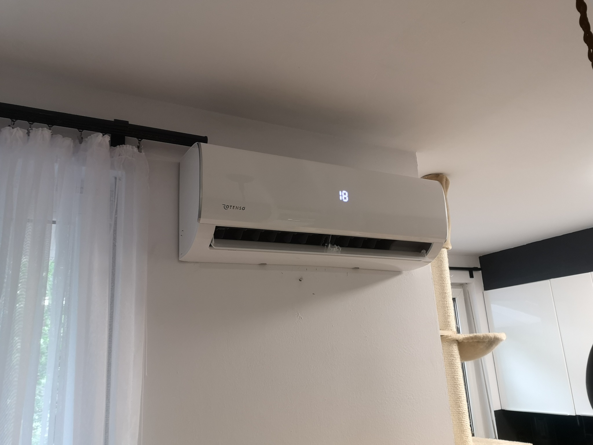 Zamontowana jednostka klimatyzacji Rotenso na białej ścianie w mieszkaniu, z widocznym wyświetlaczem temperatury 18 stopni Celsjusza, obok okna z firanką i drapakiem dla kota.