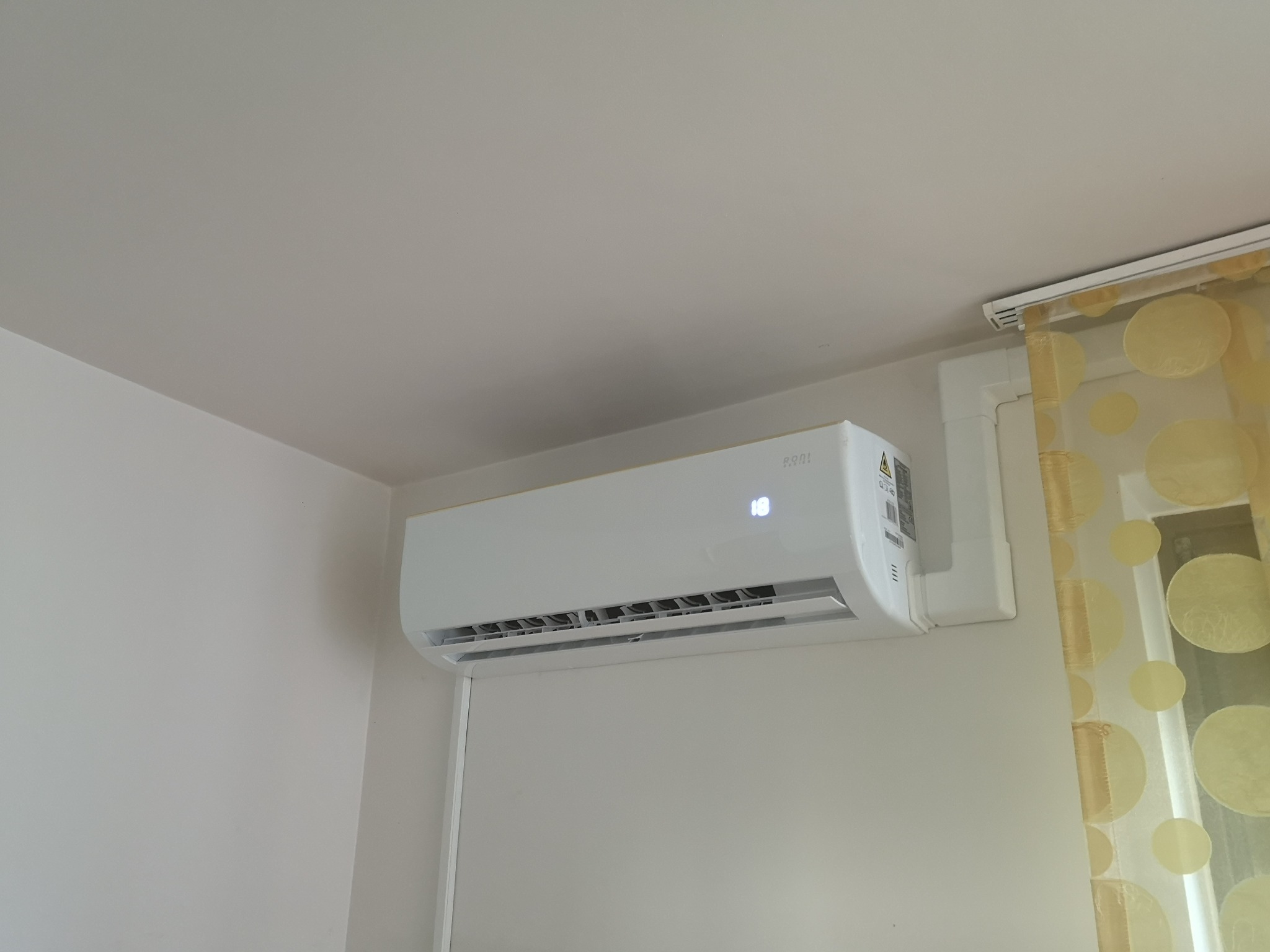 Biały klimatyzator ścienny z wyświetlaczem temperatury '18' zamontowany w pomieszczeniu z jasnymi ścianami i zasłonami w żółte grochy.