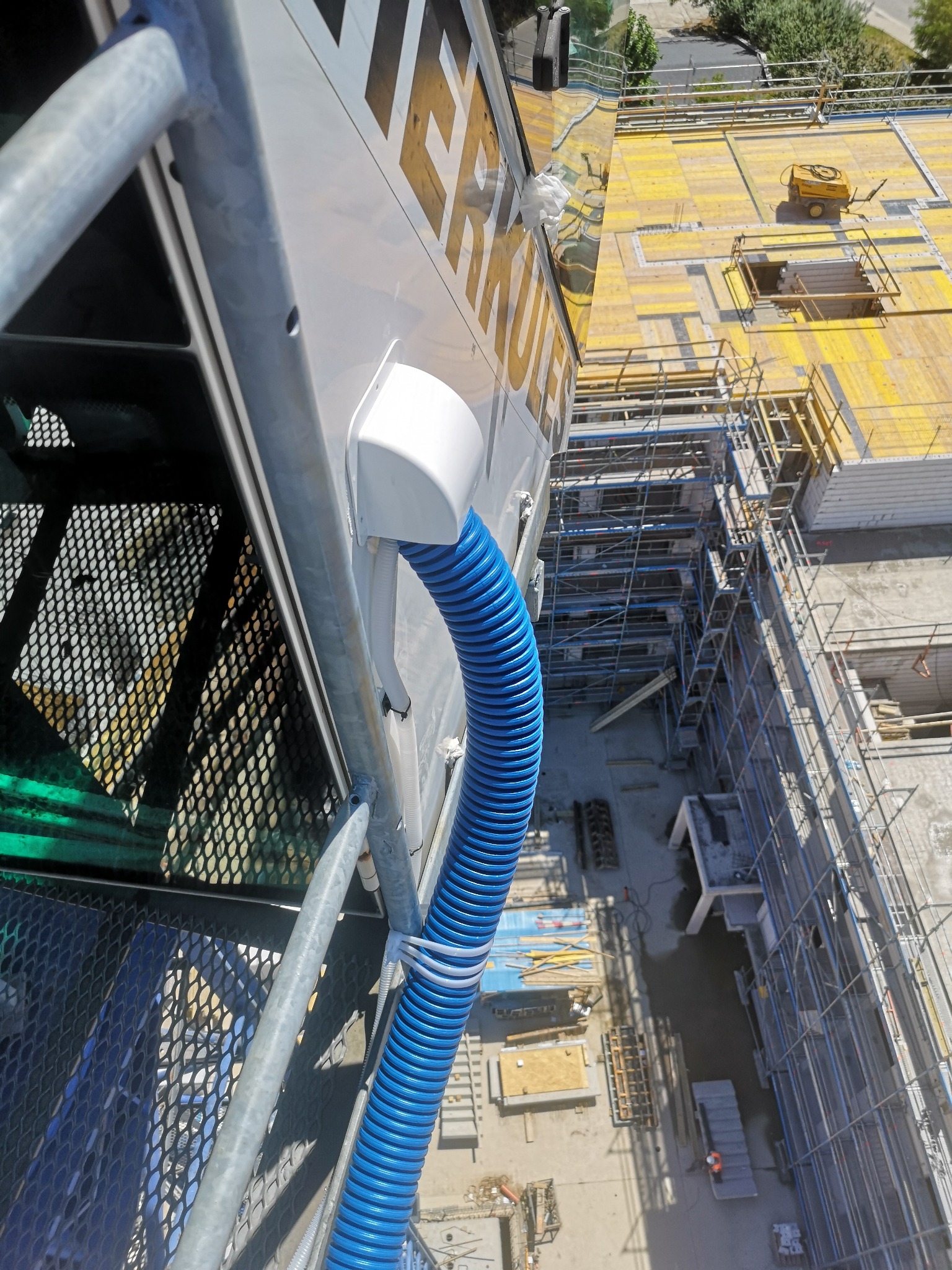 Niebieski, karbowany przewód wentylacyjny przymocowany do boku białej obudowy urządzenia na placu budowy, widok z góry na rusztowania i żółte płyty szalunkowe.