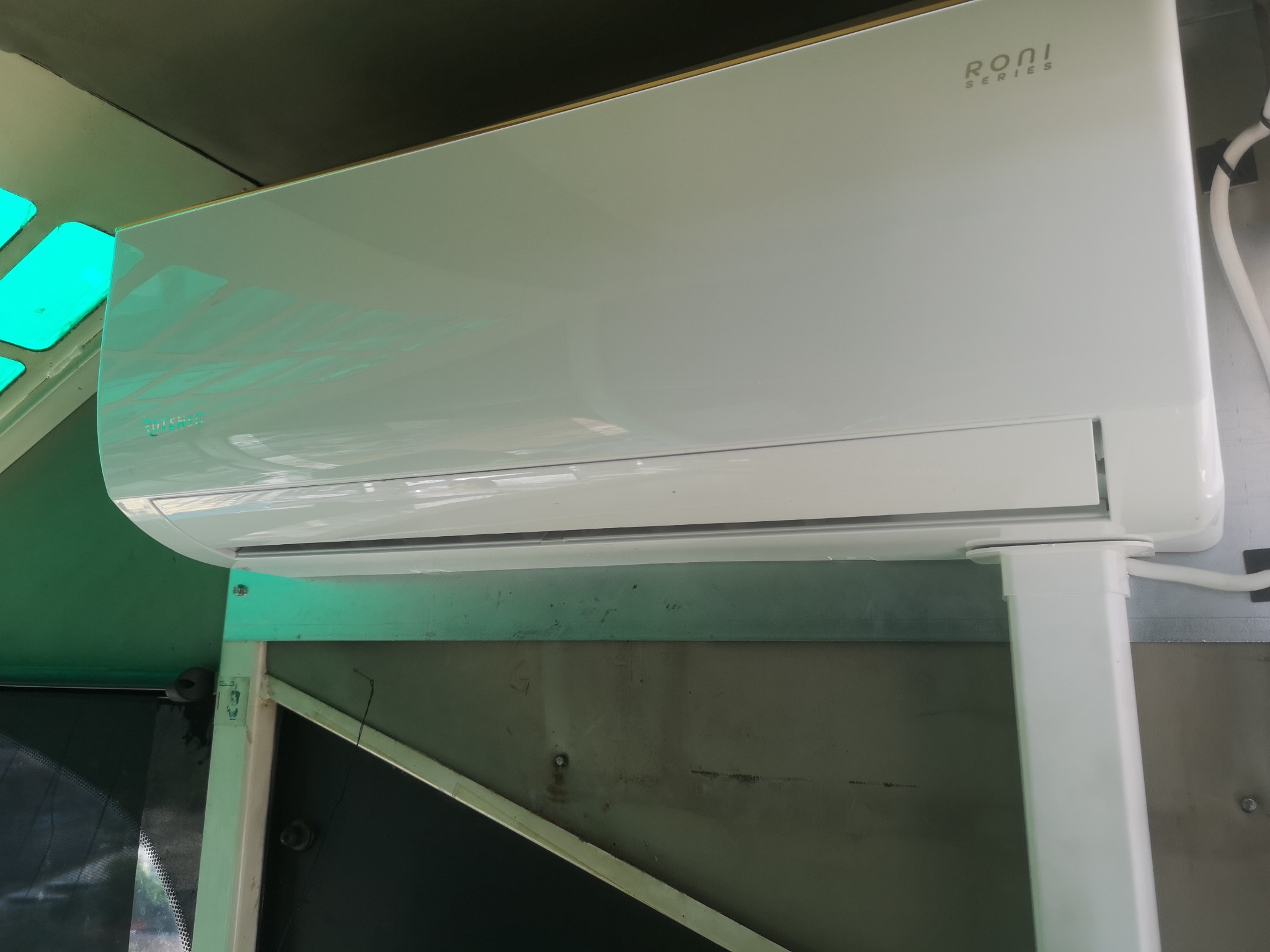 Biała jednostka klimatyzacji Roni Series zamontowana na metalowej konstrukcji z widocznymi śrubami i przewodami, oświetlona zielonym światłem.