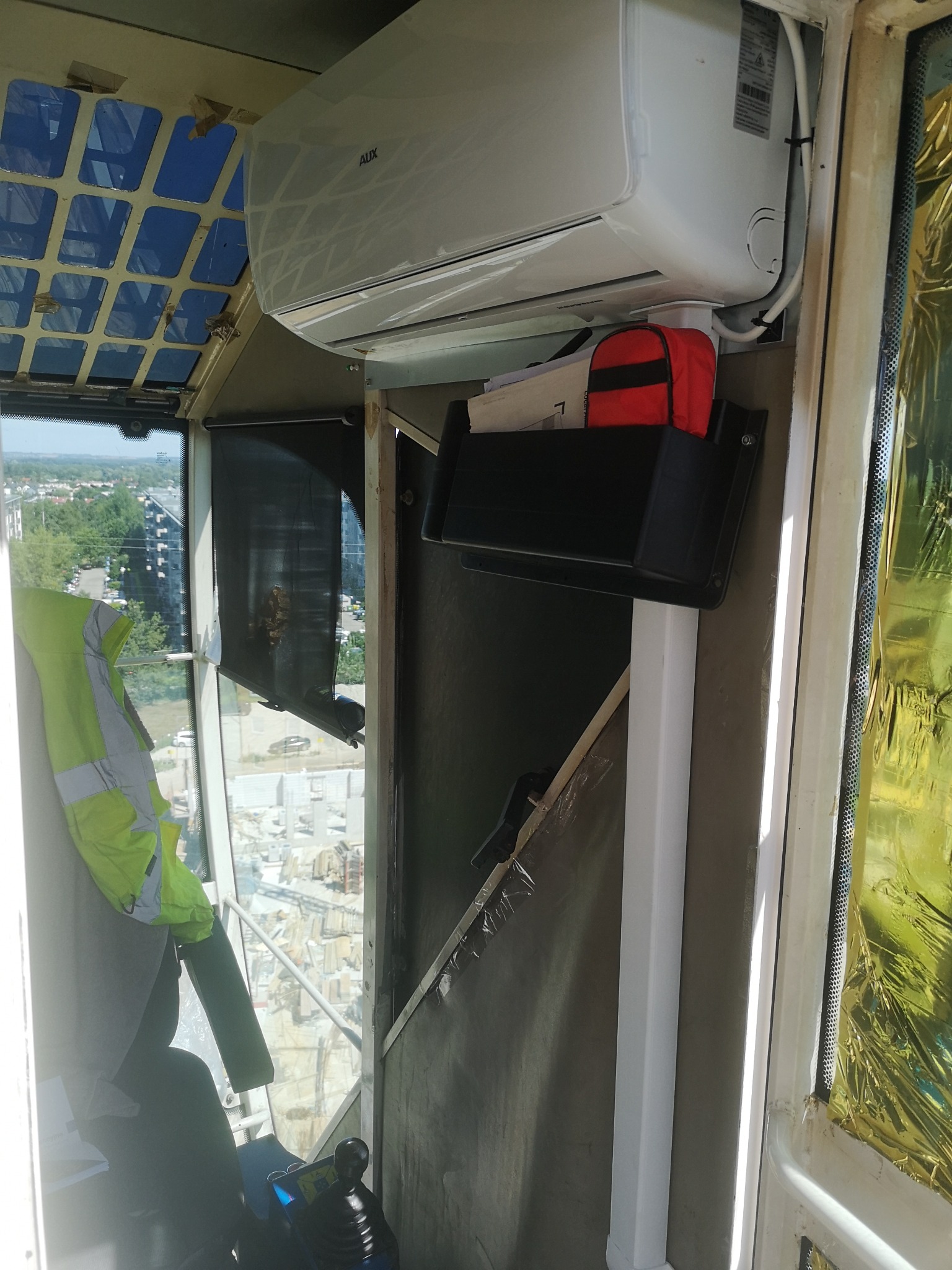 Wnętrze kabiny dźwigu z zamontowaną klimatyzacją AUX, widok na miasto w tle, kamizelka odblaskowa zawieszona na ścianie.