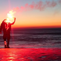 Dominik Iwon - Sylwetka osoby z flarą na tle zachodzącego słońca nad morzem. Czerwona poświata oświetla brzeg, tworząc dramatyczny efekt wizualny. Ujęcie z dołu.