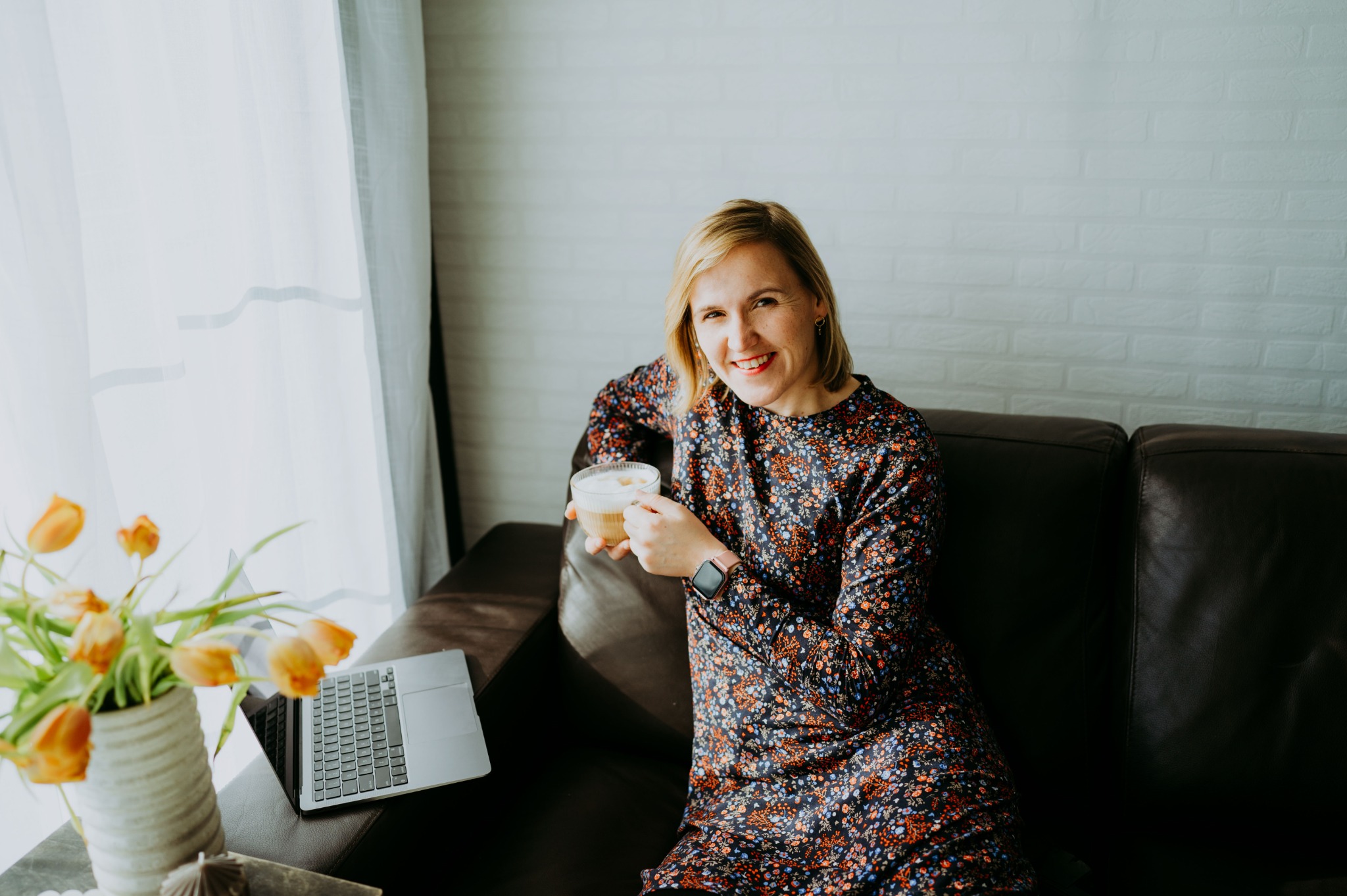 Uśmiechnięta kobieta w kwiecistej sukience siedzi na skórzanej kanapie z laptopem i tulipanami, trzymając szklankę latte, w tle biała ceglana ściana i zasłonięte okno.