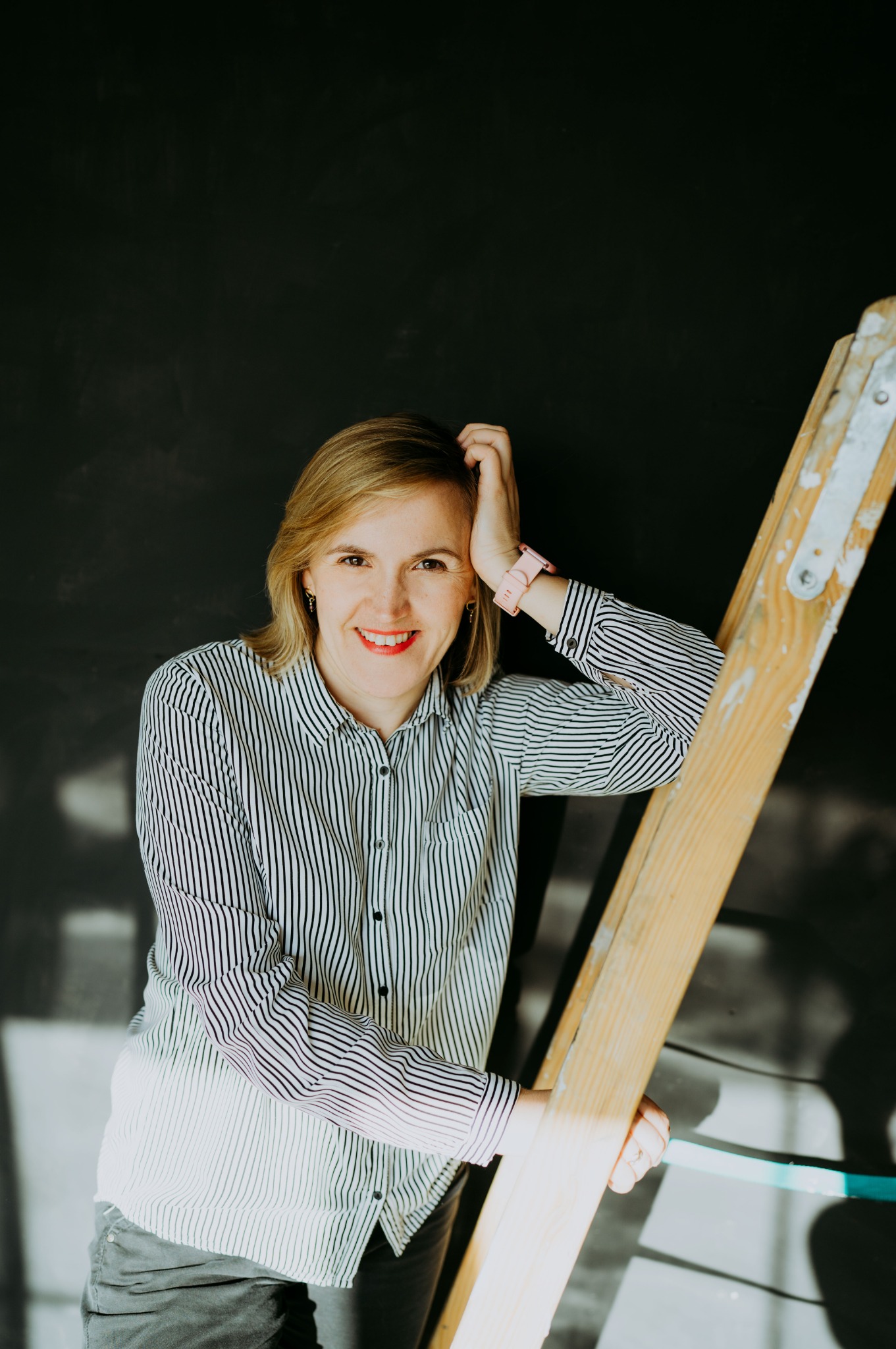 Uśmiechnięta kobieta w bluzce w paski opiera się o drewnianą drabinę na tle ciemnej ściany, prezentując profesjonalny wizerunek.
