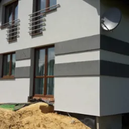 Nowoczesna elewacja domu jednorodzinnego z poziomymi pasami w odcieniach szarości, widoczna antena satelitarna, teren wokół domu w trakcie prac wykończeniowych z widoczną stertą piasku i samochodem...