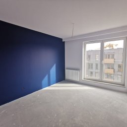 Tomasz Błaszczykowski - Wnętrze pokoju w trakcie remontu: jedna ściana pomalowana na granatowo, pozostałe białe, szara podłoga, grzejnik pod oknem z widokiem na budynek w budowie.