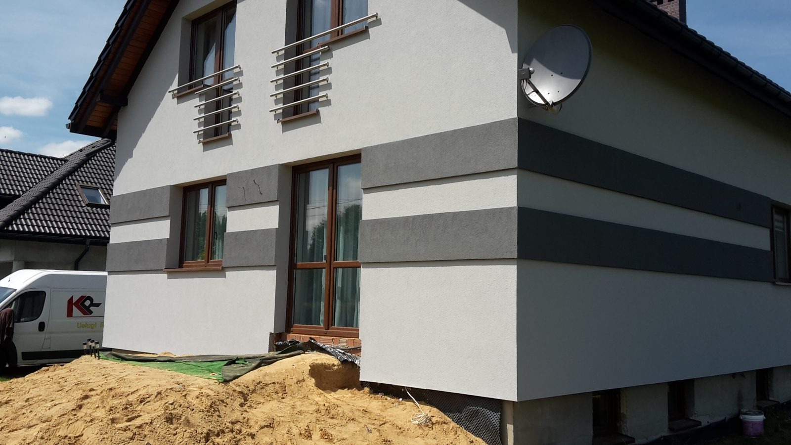 Nowoczesna elewacja domu jednorodzinnego z poziomymi pasami w odcieniach szarości, widoczna antena satelitarna, teren wokół domu w trakcie prac wykończeniowych z widoczną stertą piasku i samochodem...