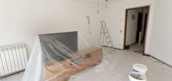 Pokój w trakcie odświeżania: świeżo pomalowane białe ściany, meble zabezpieczone folią, drabina, pędzle i wiaderka z farbą na podłodze. Widoczne niedoskonałości malarskie przy futrynie drzwi.
