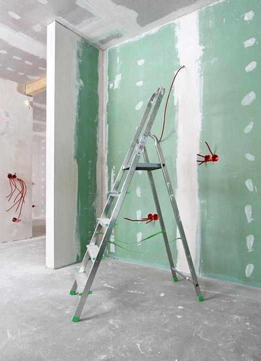 Wnętrze w trakcie prac przygotowawczych, ściany z płyt gipsowo-kartonowych częściowo pokryte zielonym podkładem, widoczne puszki elektryczne z wystającymi przewodami, aluminiowa drabina z zielonymi...