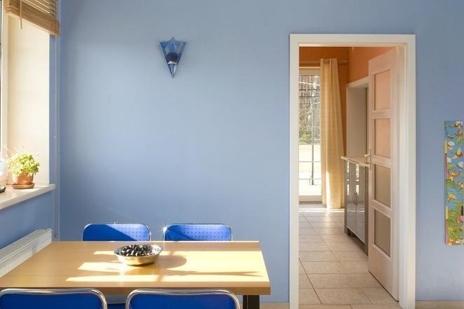 Jasnoniebieski pokój z drewnianym stołem, niebieskimi krzesłami i misą z owocami, widok na otwarte drzwi do innego pokoju z beżowymi zasłonami.